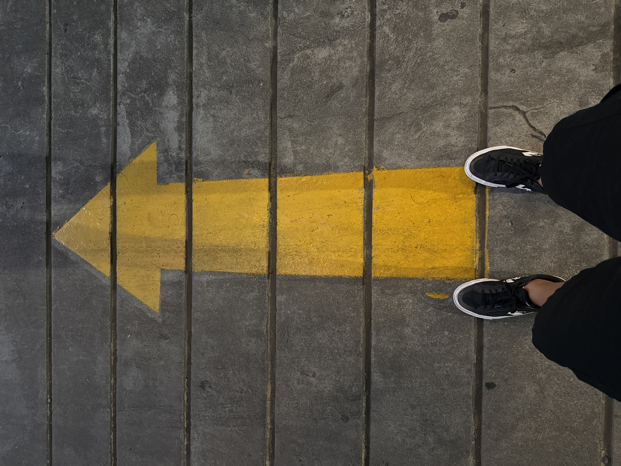 The image shows a top-down view of a person's feet wearing black sneakers with white soles, standing on a grey concrete ground marked with a bold yellow arrow pointing to the left. The concrete is composed of vertical slabs with visible grooves between them. The yellow arrow is painted directly onto the concrete surface, with some wear and tear indicating it has been there for some time. The person is wearing black pants. The lighting suggests it is daytime, possibly overcast, or the scene is in a shaded area, as there are no strong shadows. The setting appears to be an outdoor public space, such as a sidewalk or a pathway, in Nha Trang Ward, Vietnam. There is no visible text in the image. The composition focuses on the interaction between the person's choice of direction, indicated by the arrow, and their current stance.