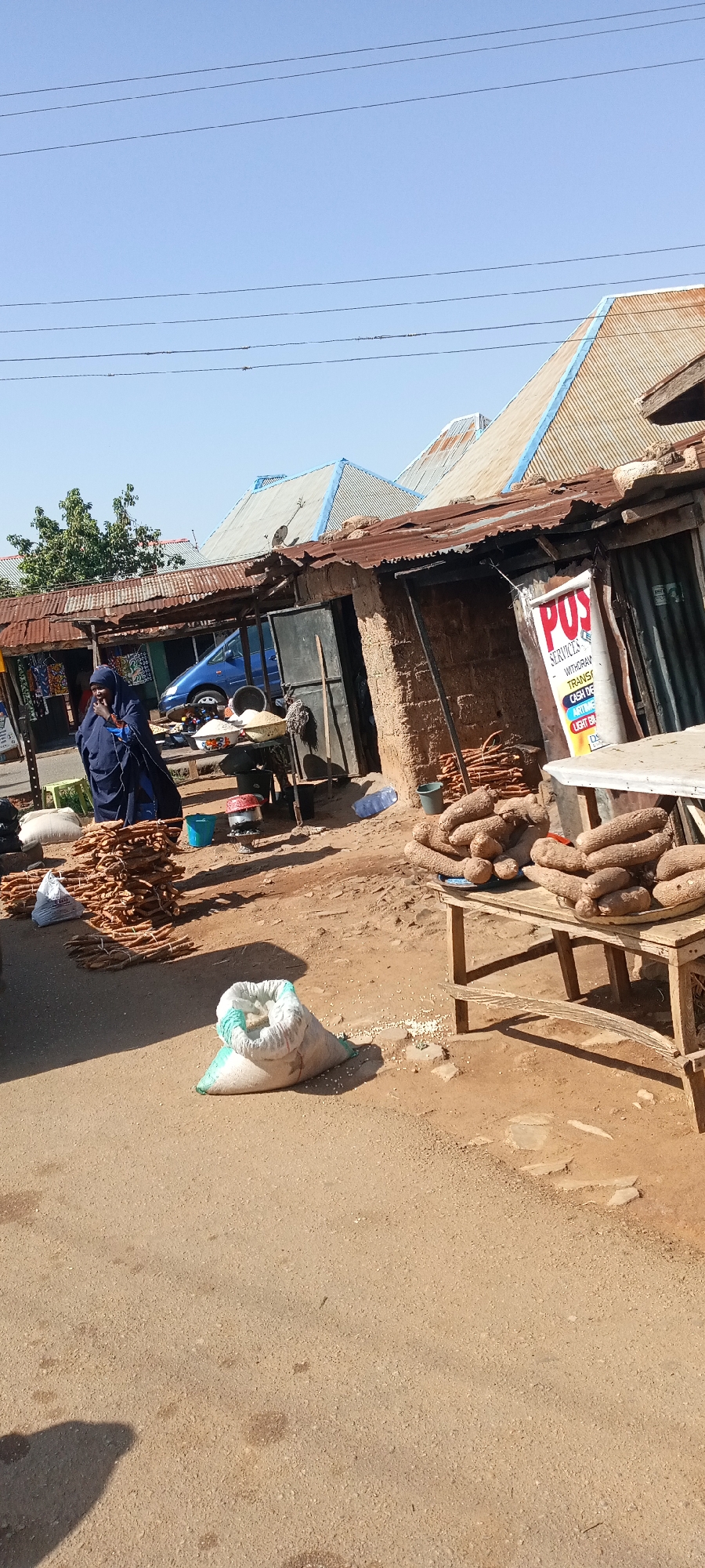 The image shows a street scene in Jos, Nigeria. A woman in a dark blue garment stands next to stacks of yam tubers. A table displays more yams. A white sack rests on the ground. In the background, a stall has a sign that reads "POS Services". The surrounding buildings have corrugated metal roofs. A blue car is partially visible. Overhead, electrical wires are strung. The environment appears dry and dusty.