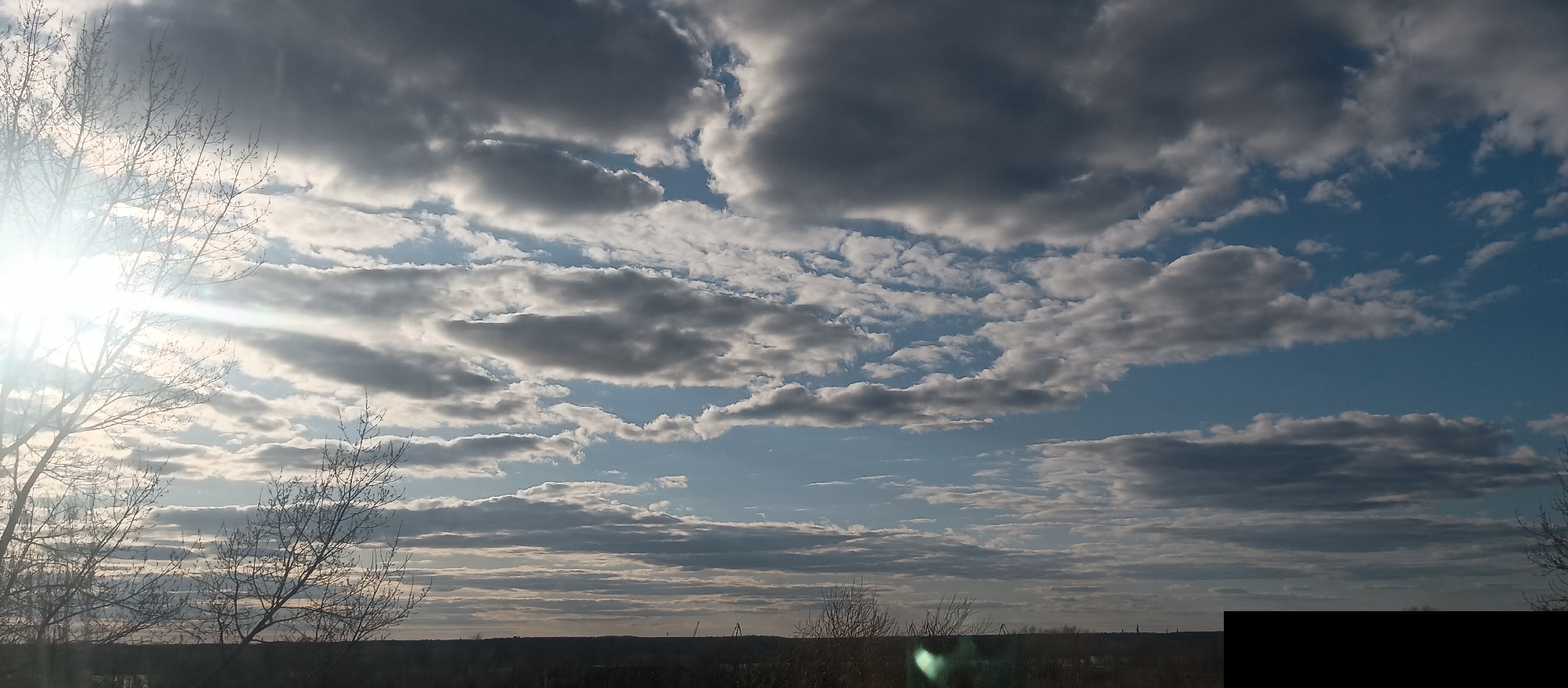 The image shows a view of the sky with layered clouds in shades of gray and white, contrasting with patches of blue. Sunlight is visible on the left side, with rays streaking across the frame. Bare branches of trees are silhouetted in the lower left and right corners. The horizon line is indistinct, with darker areas suggesting distant trees and buildings. The location is Unknown, Unknown.