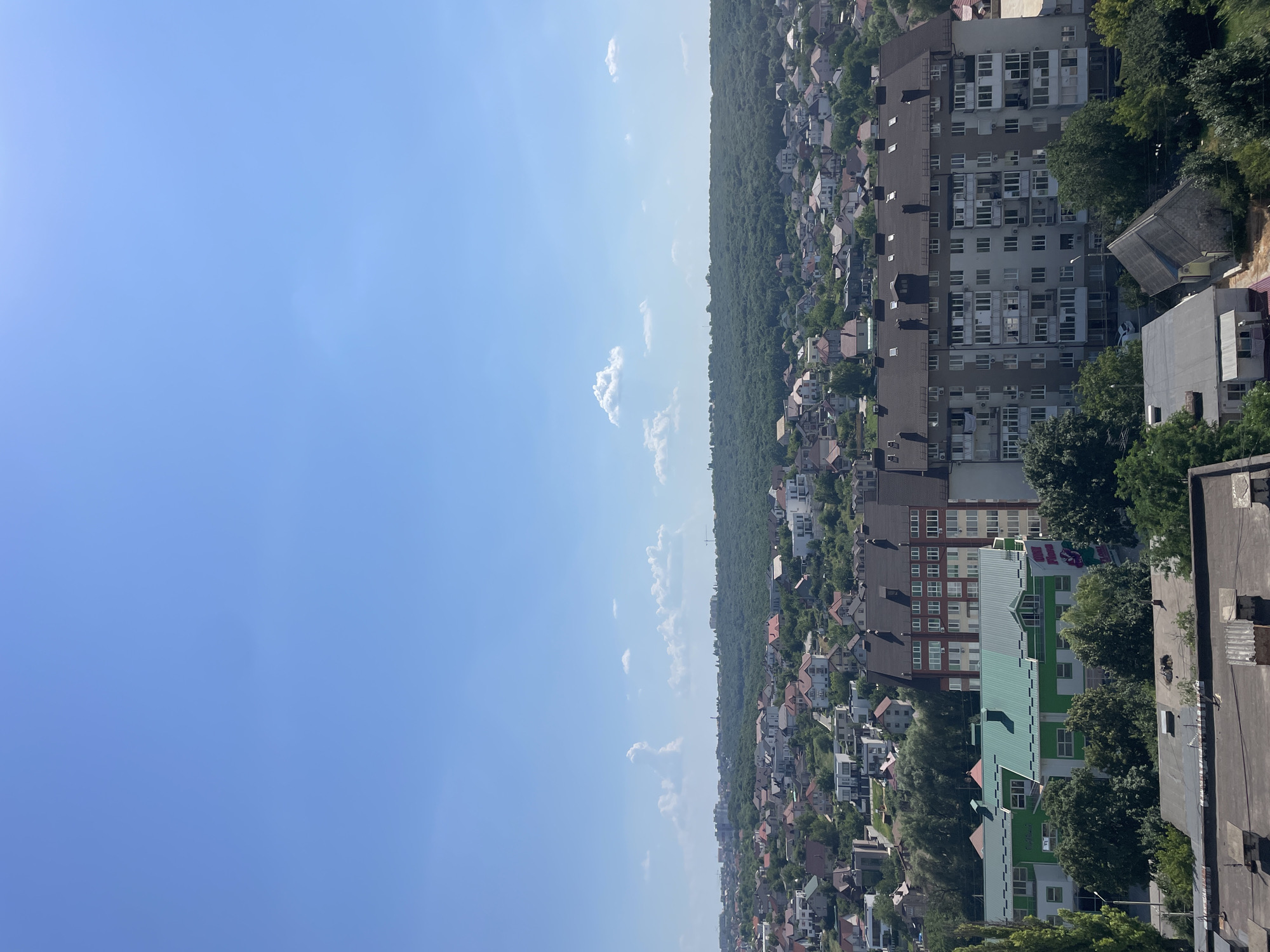 An aerial view captures a sprawling cityscape under a clear blue sky dotted with sparse, fluffy white clouds. In the foreground, flat-roofed buildings with visible chimneys suggest an urban or suburban environment. The midground is dominated by a dense collection of residential buildings, varying in height and architectural style, interspersed with lush green trees. A prominent multi-story apartment building with a dark brown roof and a facade of beige and brown panels is centrally located. To its left, a lower building features a vibrant green roof and white walls with some signage. Further back, a vast expanse of dense forest stretches across the horizon, its dark green canopy creating a striking contrast with the lighter tones of the city. The overall impression is one of a well-established residential area nestled against a significant natural landscape.