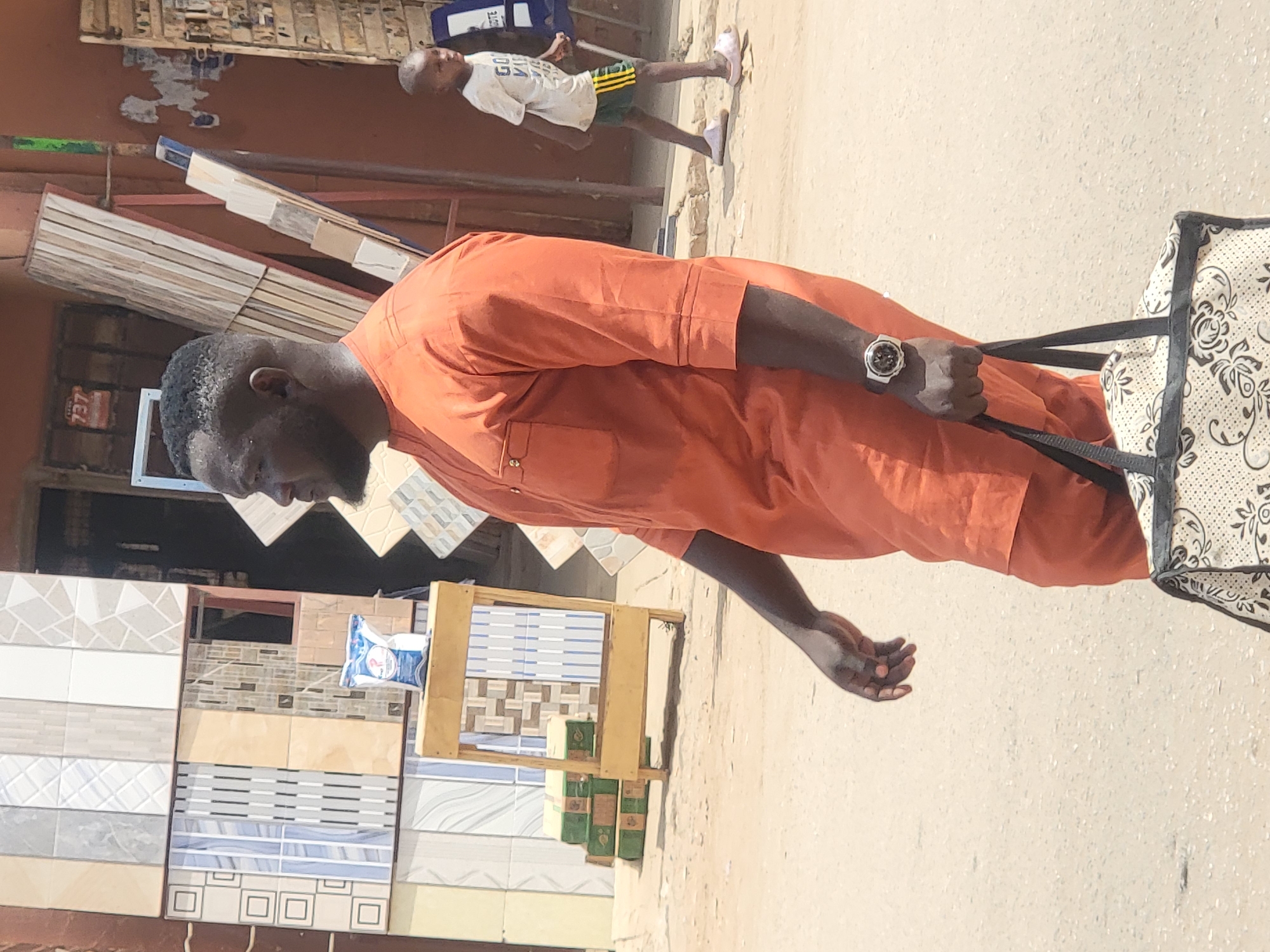 In Maiburiji, Nigeria, an adult male wearing an orange traditional outfit and a wristwatch holds the straps of a beige and black patterned bag. To his left are displays of various tiles. In the background, a child wearing a white shirt and dark shorts walks away from the main subject. The setting appears to be an outdoor marketplace or commercial area.