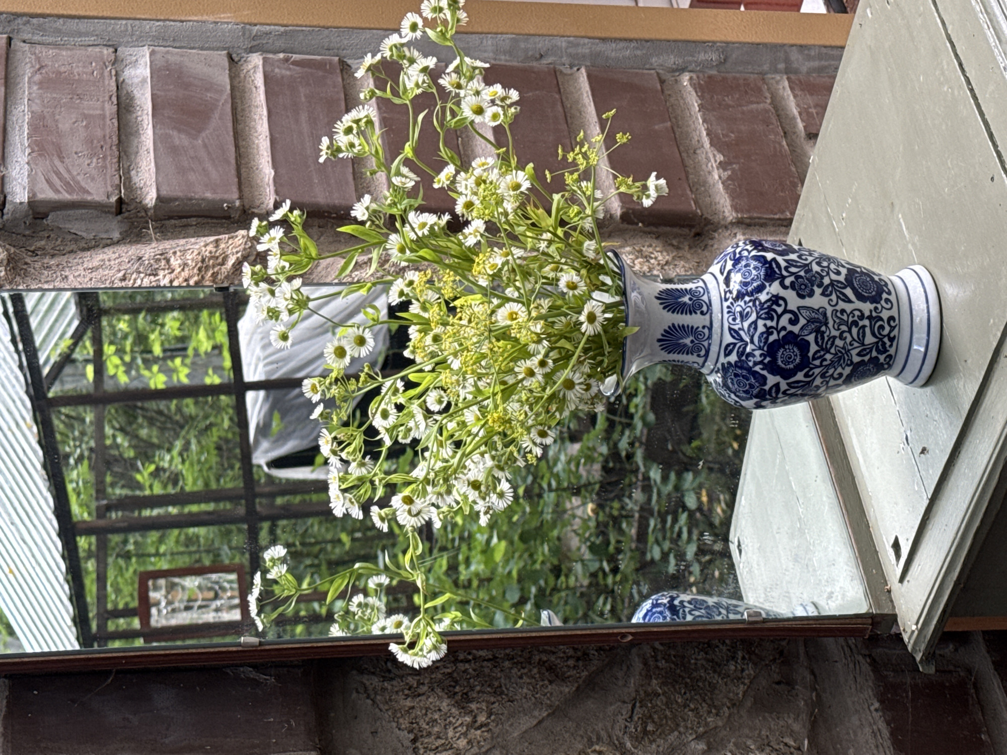 A close-up, eye-level shot focuses on a bouquet of small white flowers with yellow centers, arranged in a blue and white porcelain vase. The vase is placed on a light green wooden surface. To the left of the vase, a mirror reflects a garden scene with lush greenery and a greenhouse structure. The background features a brick wall and a rough-textured stone wall. The image does not contain any people or depict any actions or interactions. There is no discernible information to suggest the location is in the city of Perm, Russia.