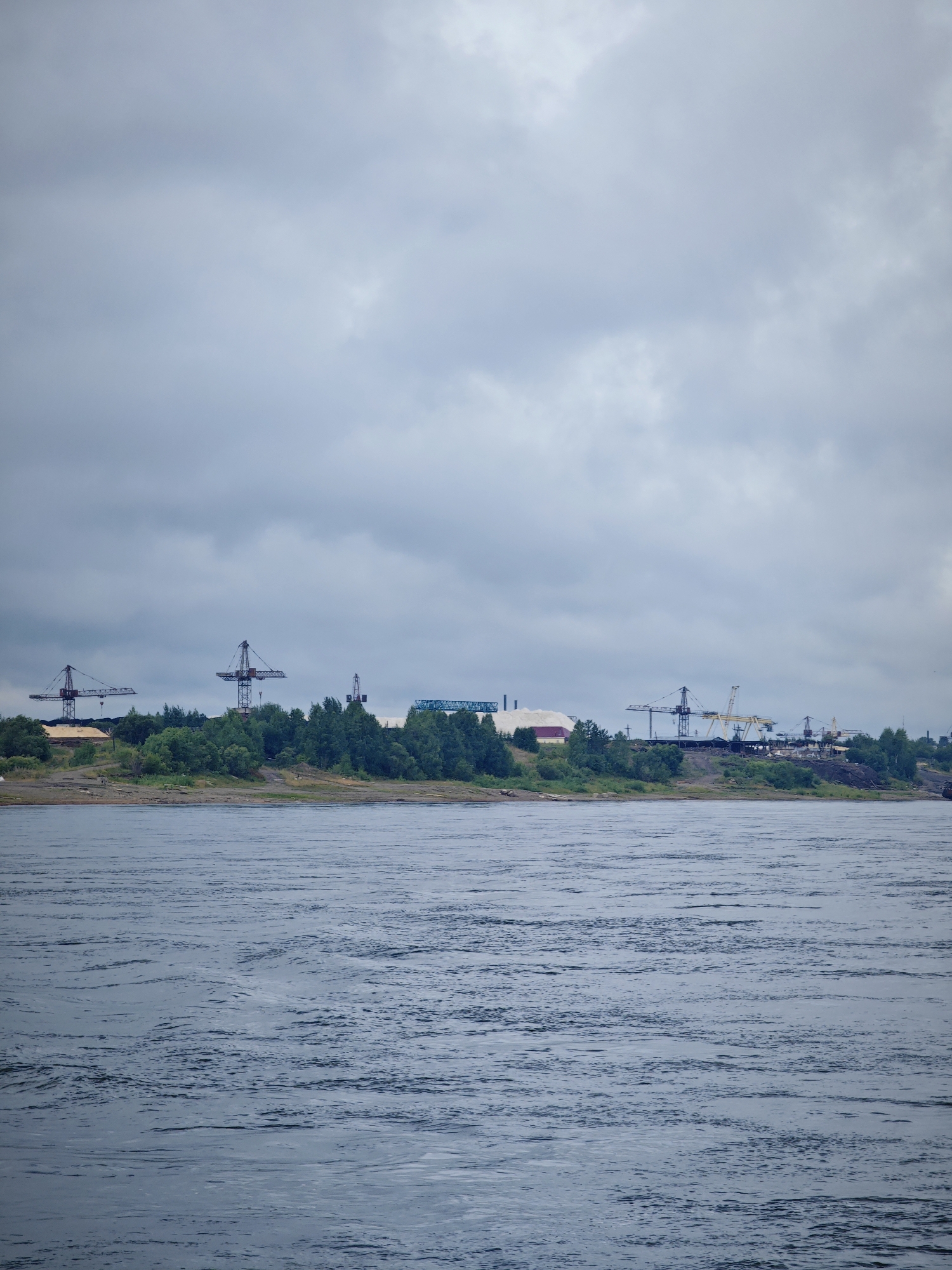 A wide, choppy river flows in the foreground under a cloudy, overcast sky. In the middle ground, a tree-lined bank separates the river from a distant industrial area. The industrial area features several large, tower cranes, some with visible boom structures, and various buildings, including one with a white, sloped roof and another with a light-colored facade. White piles are visible on the left side of the industrial area, suggesting a bulk material storage facility. The scene appears to be a port or industrial complex on the Yenisei River, likely within or near the city of Lesosibirsk, Russia, given the presence of such infrastructure and the characteristic wide river. No people are visible in the image.
