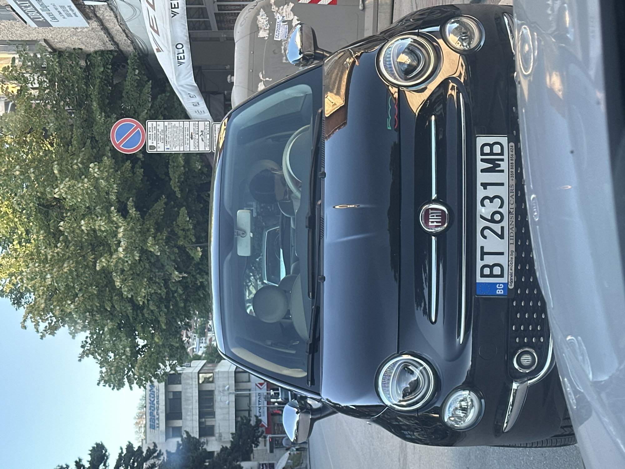 The image shows a dark-colored Fiat 500 parked on a street in Veliko Tarnovo, Bulgaria. The car has a Bulgarian license plate "BT 2631 MB." The front of the car, including the Fiat logo and headlights, is prominently displayed. A "no stopping" sign is visible in the background. The "Булстрад" building is visible across the street. A sign for "Адвокат Скалея Шарков" (Lawyer Skaleya Sharkov) can also be seen in the background.