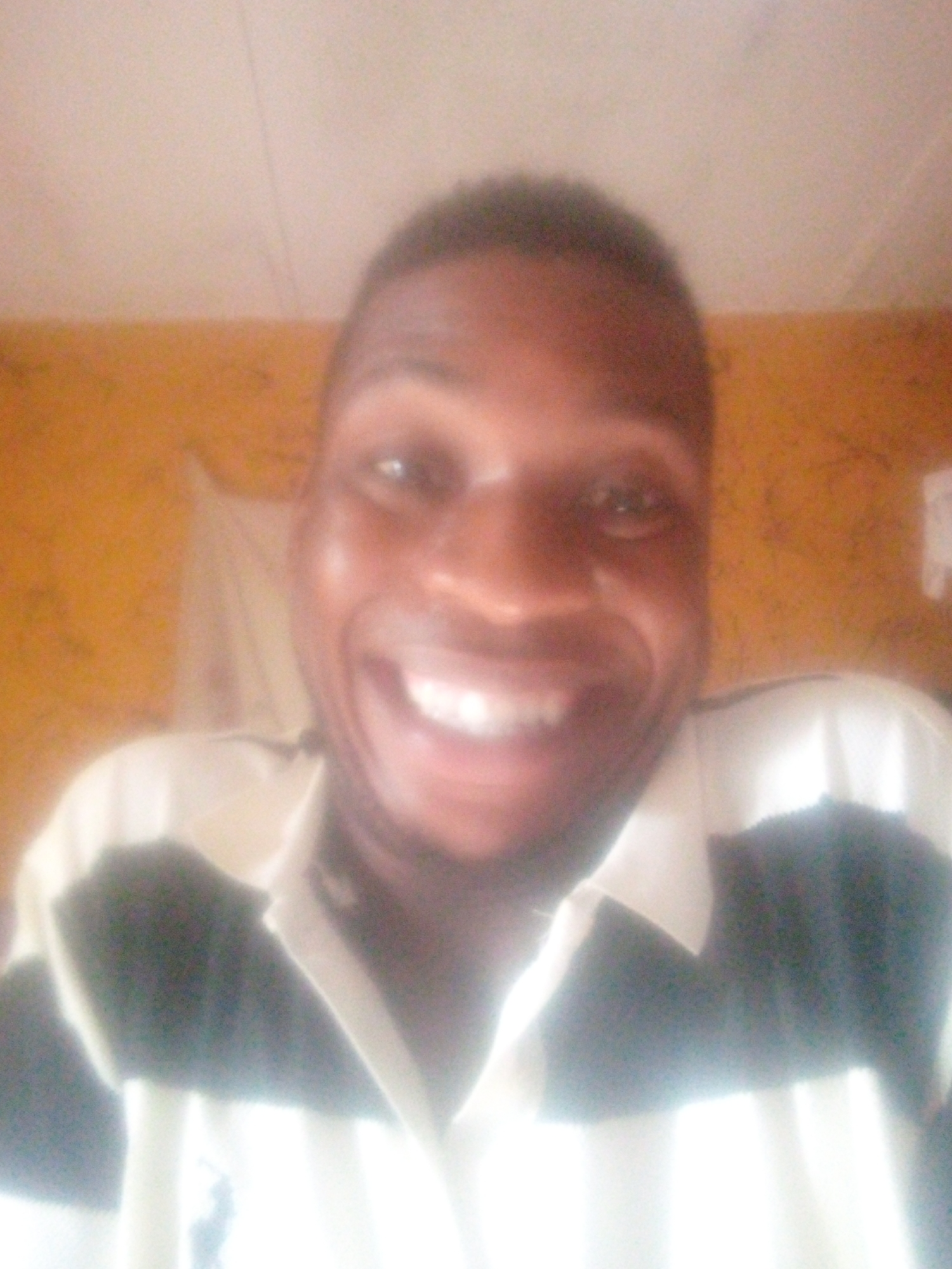A close-up shot of a man with dark skin and short, dark hair is shown smiling widely. He is wearing a black and white striped collared shirt. The background is blurred, but appears to be an interior setting with yellow-toned walls. The location is unknown.