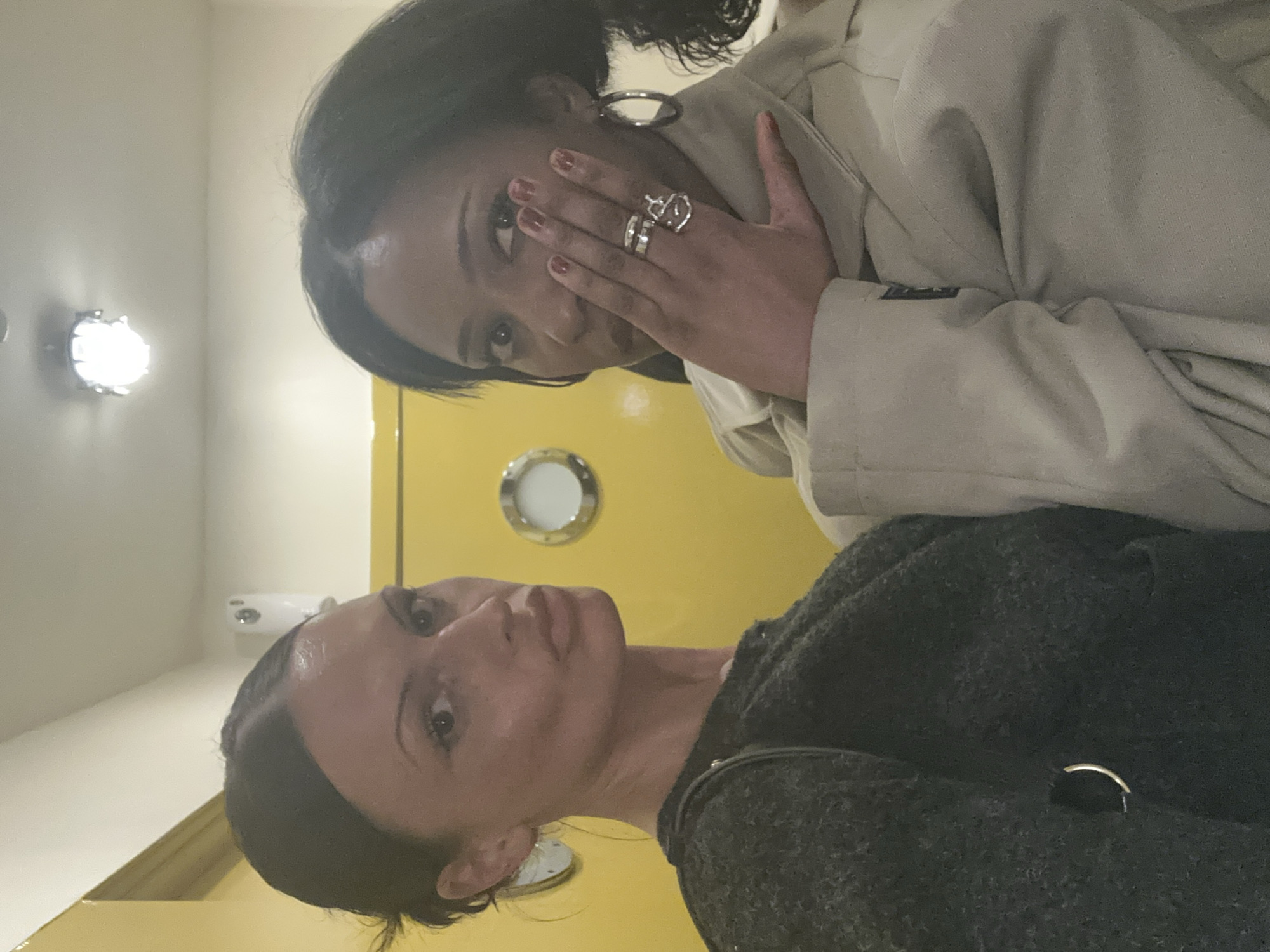 Two women are photographed indoors against a yellow wall with a porthole-style window. The top woman has dark hair, wears large hoop earrings, multiple rings, and a light-colored trench coat. She holds her hand up to her face, partially covering her eye. The lower woman has dark hair pulled back from her face and wears a dark grey sweater. The photo appears to be taken at an upward angle. Based on the location in London, it is possible they are in a themed restaurant, bar, or tourist attraction with a nautical theme.