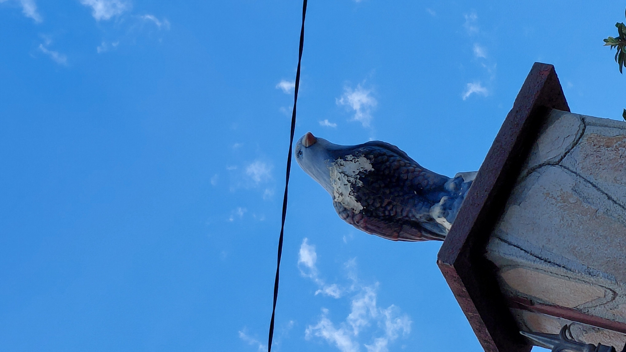A low-angle shot depicts a ceramic eagle perched atop a weathered structure against a clear blue sky dotted with scattered white clouds. A thin, black electrical wire bisects the upper left quadrant of the frame. The eagle is predominantly gray and brown, with chipped paint revealing underlying layers. The supporting structure appears to be made of stone or concrete with a rusty metal border. Green foliage is visible in the upper right corner, partially obscuring the sky. The location is Unknown, Unknown.