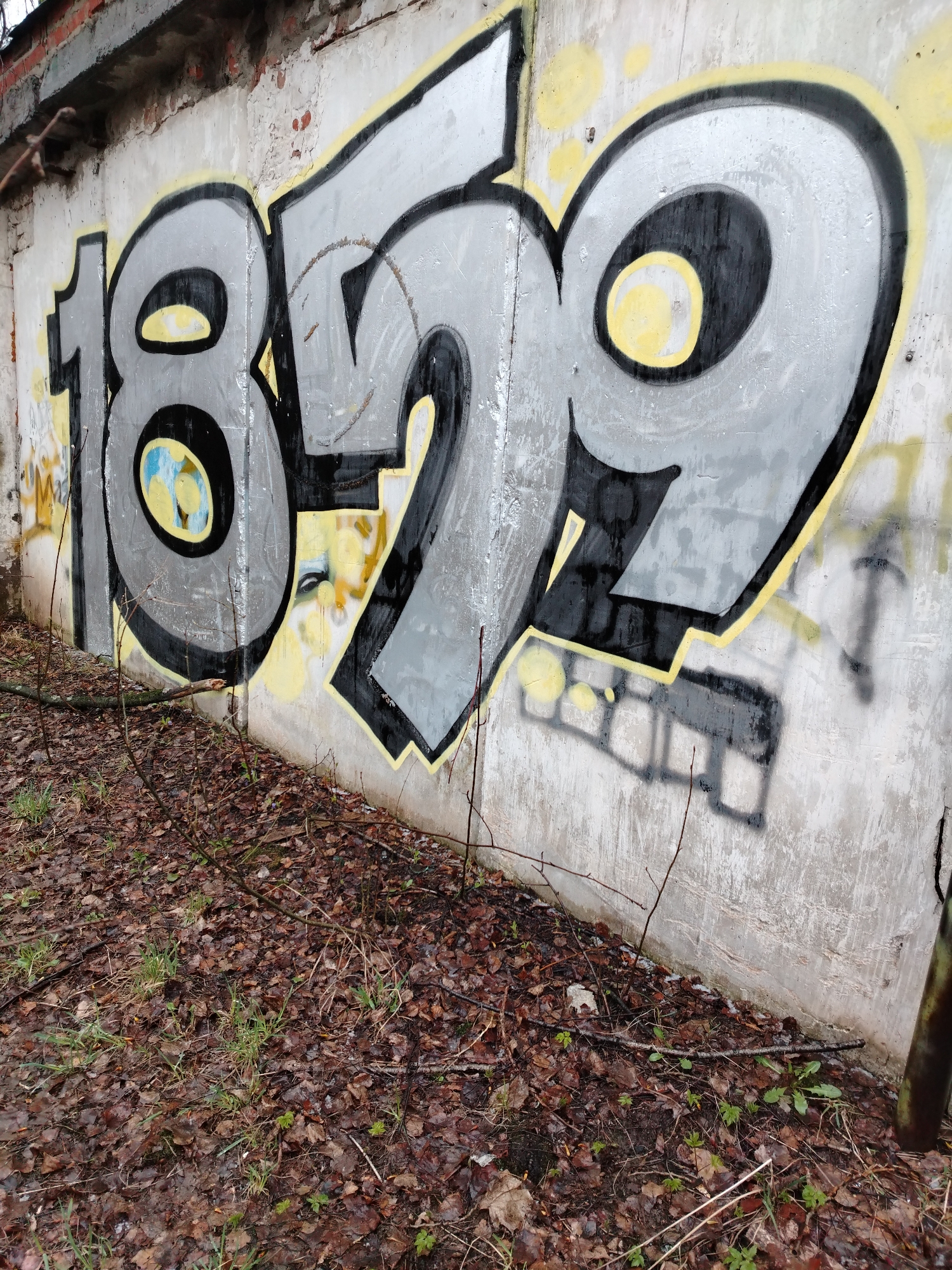 The image shows a concrete wall with graffiti, seemingly located outdoors, based on the presence of fallen leaves and sparse vegetation at the base of the wall. The graffiti consists of the number "1879" painted in a stylized, blocky font with a black outline and a silver or gray fill. Some yellow paint is present around the numbers, either as part of the graffiti or as an earlier layer. The wall itself is weathered and shows signs of wear and discoloration. The area in front of the wall appears to be covered in damp leaves and small branches. No people are visible in the image. The location is not possible to determine.
