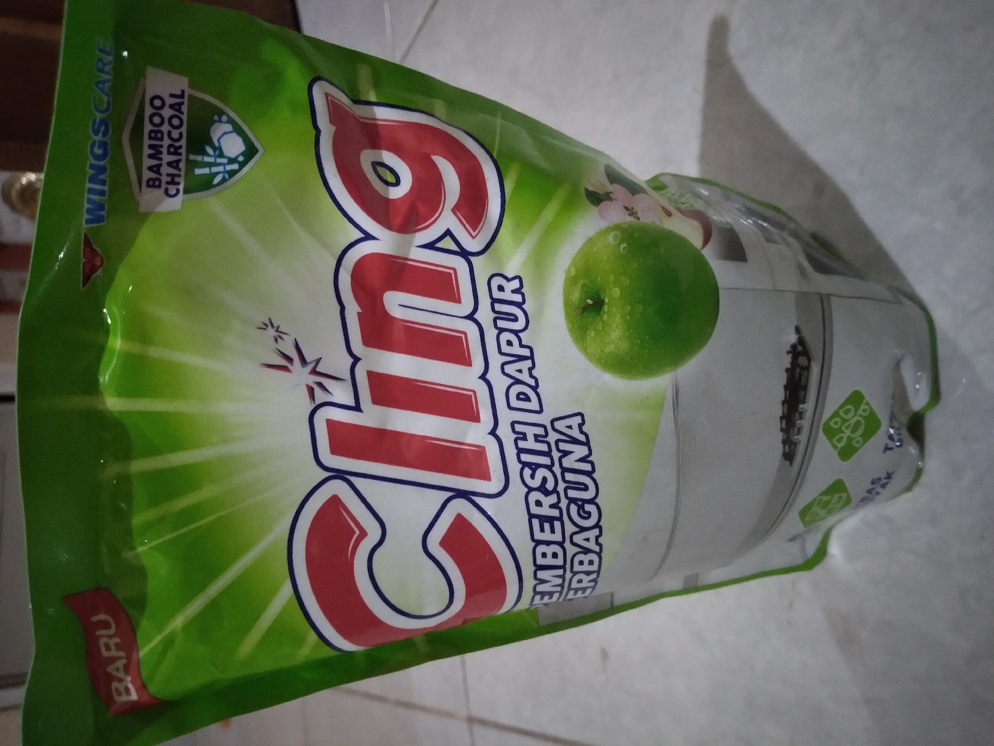 The image shows a close-up, eye-level view of a green plastic pouch of "Cling" dish soap. The pouch has a prominent "Cling" logo in red and white lettering. At the top left, there is a red banner with the word "BARU" (New) in white. To the right of "BARU", the "WINGSCARE" logo is visible in blue, followed by a shield-shaped graphic indicating "BAMBOO CHARCOAL" with an illustration of bamboo and charcoal. Below the "Cling" logo, text in blue reads "PEMBERSIH DAPUR ERBAGUNA" (Multipurpose Kitchen Cleaner). A green apple, slightly to the right of the center, is depicted on the pouch. The background appears to be a light-colored, textured surface, possibly a floor tile, with a slight shadow cast by the pouch. The scene is indoors. The image location is not specified in the provided context.