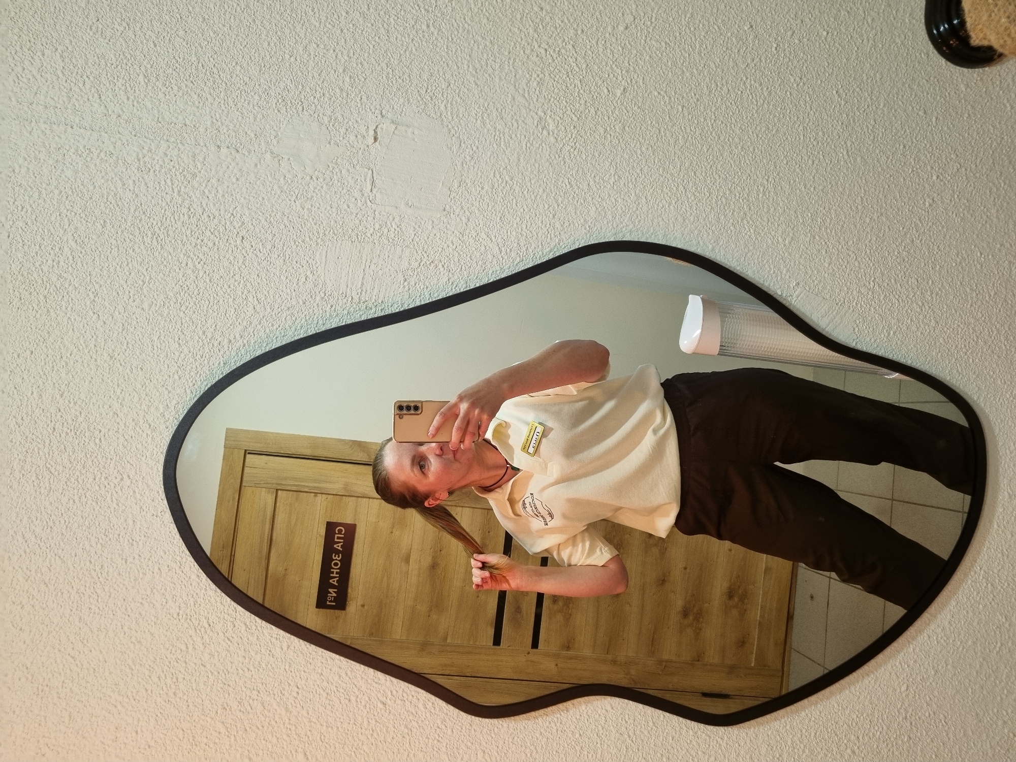 A young woman is reflected in a mirror. She is holding a phone and taking a selfie. She is wearing a light-colored polo shirt with a name tag and dark pants. The reflection in the mirror shows a wooden door with a sign in Russian that says "ГОИ ЗОНА АПС". The background also shows a wall with white tiles and a light fixture. The location is likely in Chaikovsky, Russia, given the Russian text on the sign.