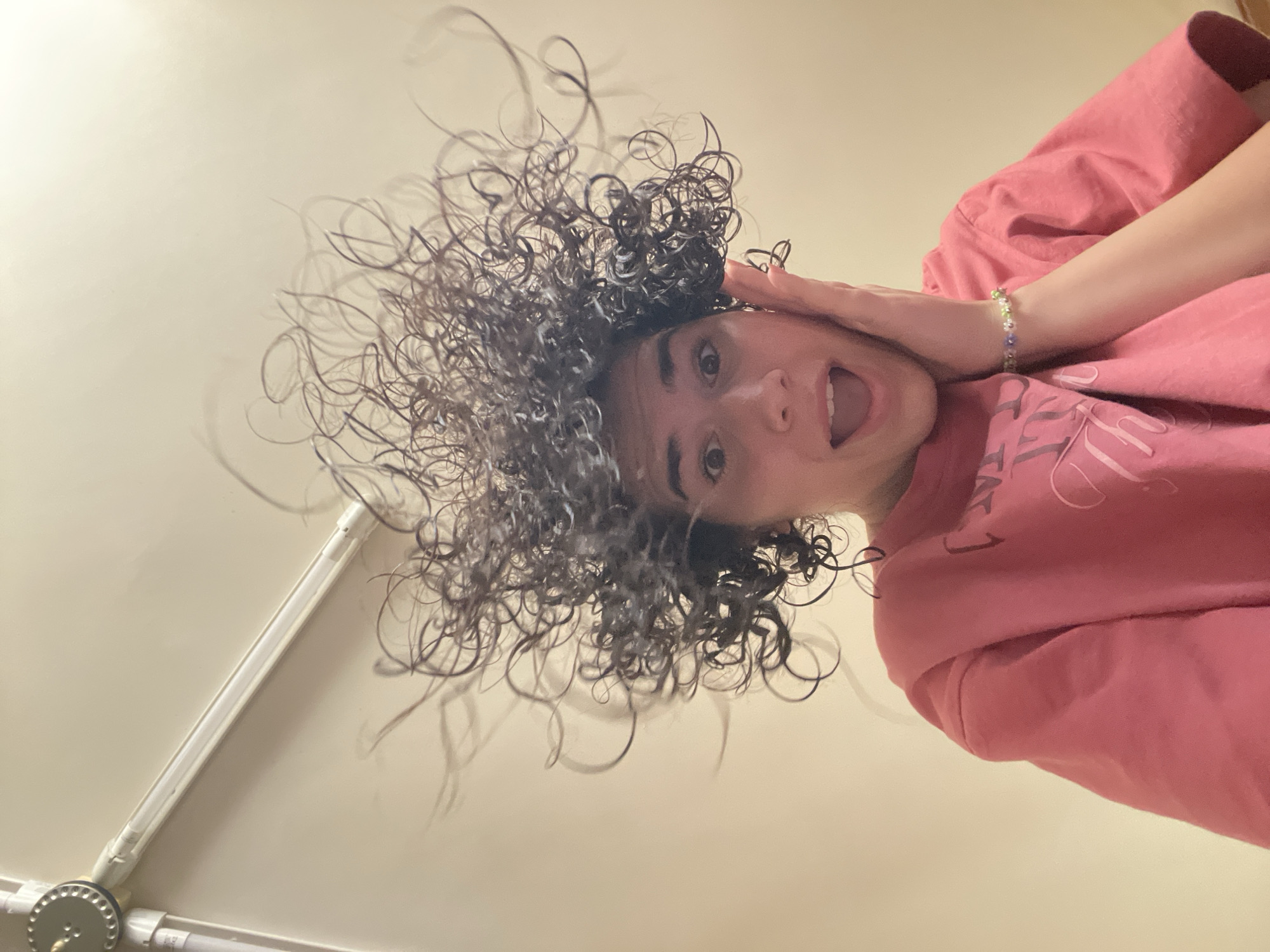 A person with curly hair and wearing a pink sweatshirt is facing downward with an open mouth, with one hand on their head and the other arm extended. A light fixture is visible on the left. The background appears to be a light-colored wall. Location: Not identifiable within Sofia, Bulgaria.