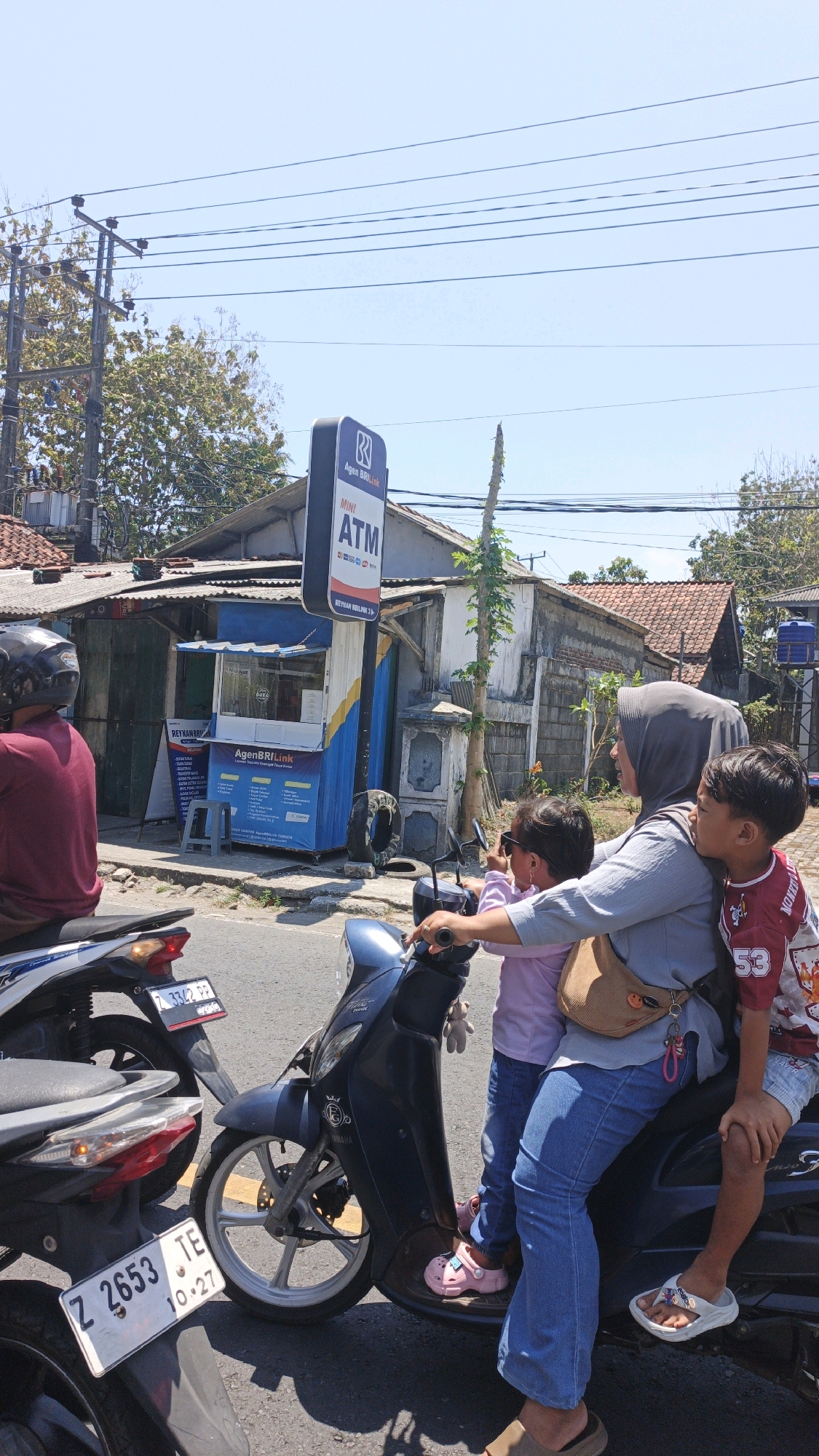 An outdoor, eye-level, medium shot captures a family on a scooter on a sunny day in Sukaresik, Indonesia. A woman wearing a grey hijab and a blue shirt sits on the scooter, holding two children. The child in front, wearing a pink shirt and pink Crocs, faces forward. The child behind, wearing a red and white shirt with the number 53, faces sideways. The scooter is dark blue. In the background, a small blue and yellow booth with an ATM sign is visible. A power pole with numerous wires stands to the left of the ATM, and trees with green foliage are seen further back. The ground is paved with asphalt. The license plate of a second scooter parked behind the family's scooter reads "Z 2653 TE 10.27".