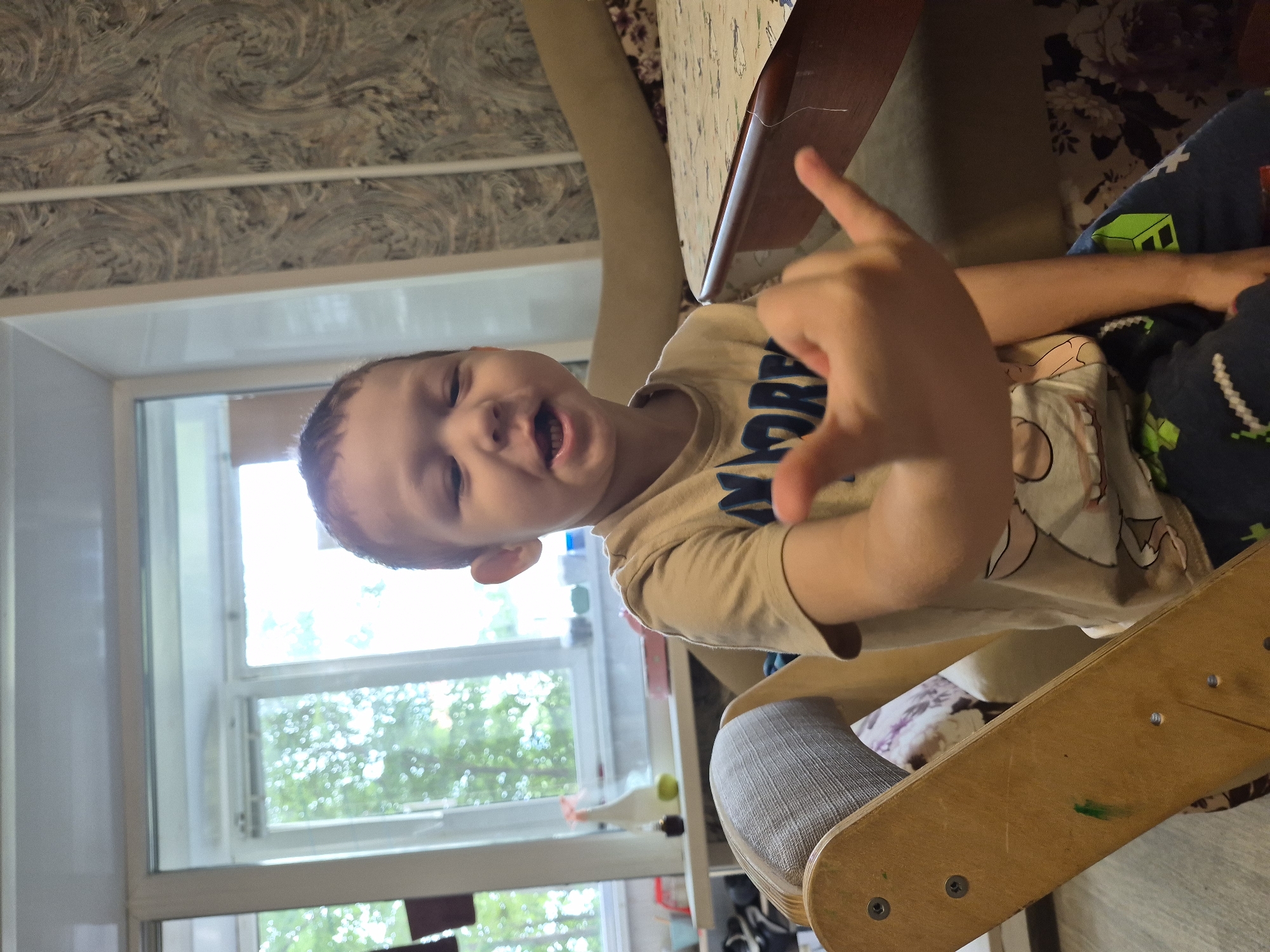 In Nyagan, Russia, a young boy is captured in a medium shot, seated on a wooden rocking toy. He is wearing a light brown t-shirt with a graphic and dark blue pants adorned with green and white pixelated designs. The boy is looking directly at the camera with his mouth slightly open, revealing his teeth. His right hand is raised, with his thumb and pinky finger extended in a gesture that resembles the "shaka" sign. The background features a window with greenery visible outside, a patterned wallpaper, and a portion of a table. The lighting suggests an indoor setting, possibly a living room.
