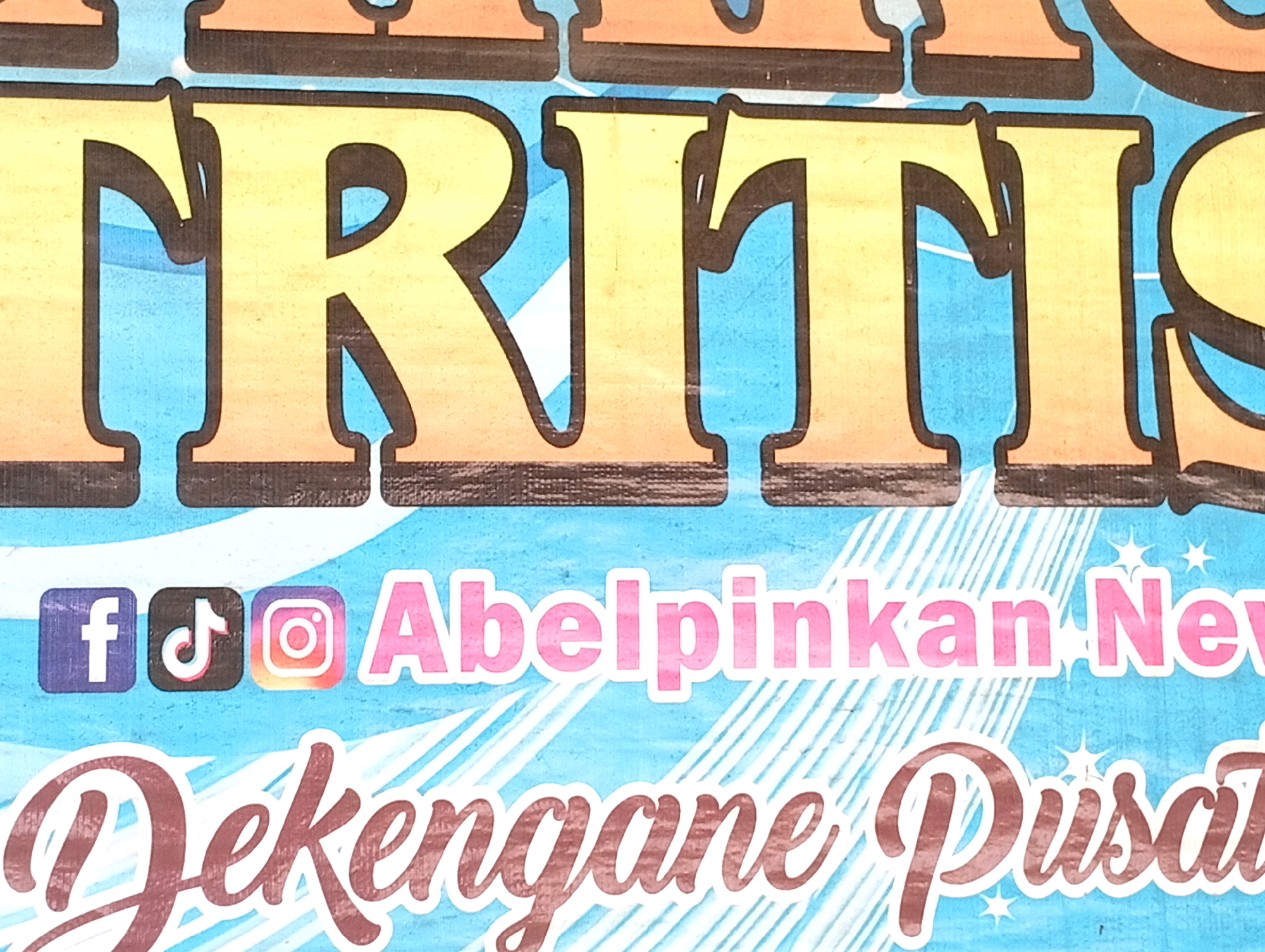This is a close-up, eye-level, horizontal, outdoor, medium shot of a colorful banner with large, bold lettering in yellow and orange with black outlines. The banner has a blue background with white wavy lines and some starbursts. In the middle of the banner, there are three social media icons: Facebook, TikTok, and Instagram, followed by pink text that reads "Abelpinkan Nev". Below this text, in a darker, cursive font with a white outline, is the word "Dekengane Pusat". The banner appears to be displayed in a public area, possibly in the city of Boyolangu, Indonesia, given the Indonesian language used in the text.