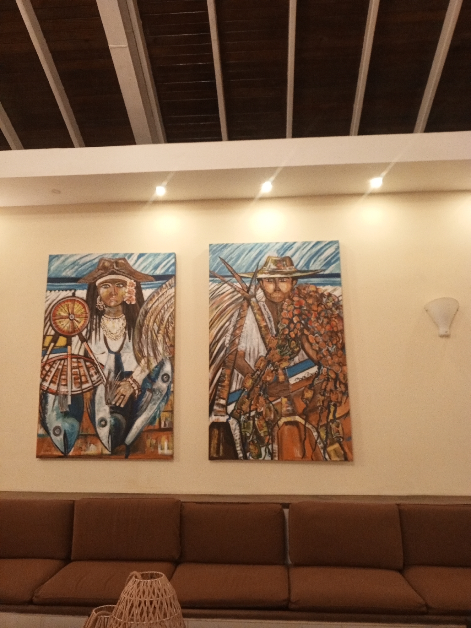 The image displays an indoor setting, likely a hotel lobby or waiting area, featuring two large, vibrant paintings on a light-colored wall. Above the wall, a white soffit houses recessed spotlights, casting illumination downwards. The ceiling above consists of dark wooden beams interspersed with lighter, perpendicular structural elements.
The two prominent artworks are rectangular and vertically oriented, depicting stylized figures associated with the sea. The painting on the left portrays a person, possibly a woman, with long dark hair, wearing a wide-brimmed hat, a necklace, and a flower near their ear. They are depicted with elements of fishing, including a large wooden wheel resembling a boat's steering wheel or fishing net component, and two large fish (possibly tuna) at the bottom. The background features blue and brown hues, suggesting a marine environment.
The painting on the right depicts another person, possibly a man with a beard, also wearing a wide-brimmed hat. This figure is adorned with or carrying a large, bountiful catch, appearing to be lobsters or crabs rendered in warm orange and red tones. Similar to the left painting, the background hints at a coastal or marine setting with blue and brown elements. Both paintings feature a folk art style, possibly representing local Venezuelan culture or traditions related to fishing in the Porlamar area.
Below the artworks, a long, brown upholstered sofa with multiple cushions provides seating. Partially visible in the foreground, in front of the sofa, is a light-colored, woven basket-like object, possibly a decorative piece or a small table. A white wall sconce is visible on the right side of the wall. The artificial lighting suggests the scene could be day or night indoors. There is no visible text.