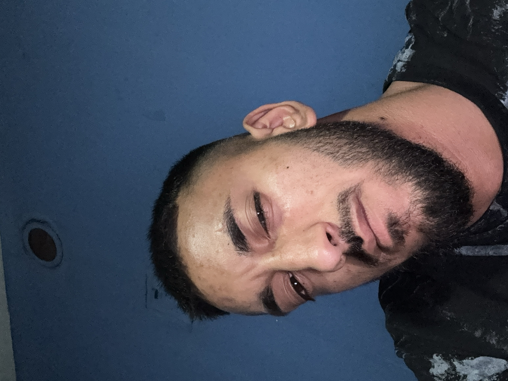 The image is a close-up selfie of a man against a blue wall. The man has short, dark hair, a beard, and is looking towards the camera with a tired expression. His right eye is partially closed. He is wearing a black t-shirt with a light-colored speckled pattern on the shoulder. The background is a plain blue wall with a circular vent or light fixture visible in the upper left corner. There is no indication of the location being in Iskritsa, Bulgaria.