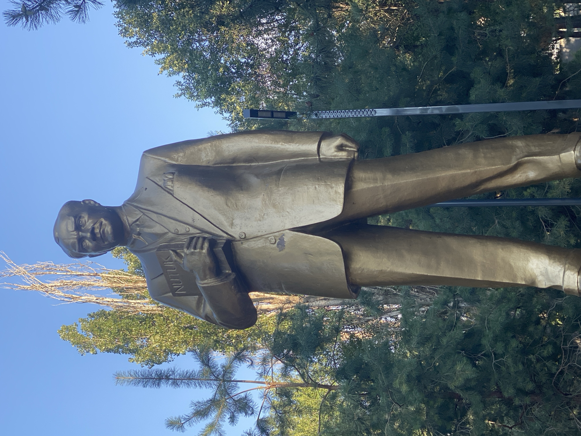A full-body bronze statue of a man wearing a suit and holding a book is depicted against a clear blue sky. The statue is positioned in the foreground, dominating the frame, with its base obscured by shadow. The man in the statue has a stern expression, his gaze directed forward. He is dressed in a suit with a jacket, vest, shirt, and tie, and he holds an open book in his left hand. The book is inscribed with the word "NUTUK."
In the background, lush green pine trees and taller, sparser deciduous trees create a natural setting. Sunlight illuminates the statue from the side, casting subtle shadows and highlighting the metallic sheen of the bronze. A modern, dark gray lamppost with a rectangular light fixture is visible to the right of the statue, its metallic structure contrasting with the organic forms of the trees and the classical statue. The overall scene suggests an outdoor public space, possibly a park or square, in Bishkek City, Kyrgyzstan.