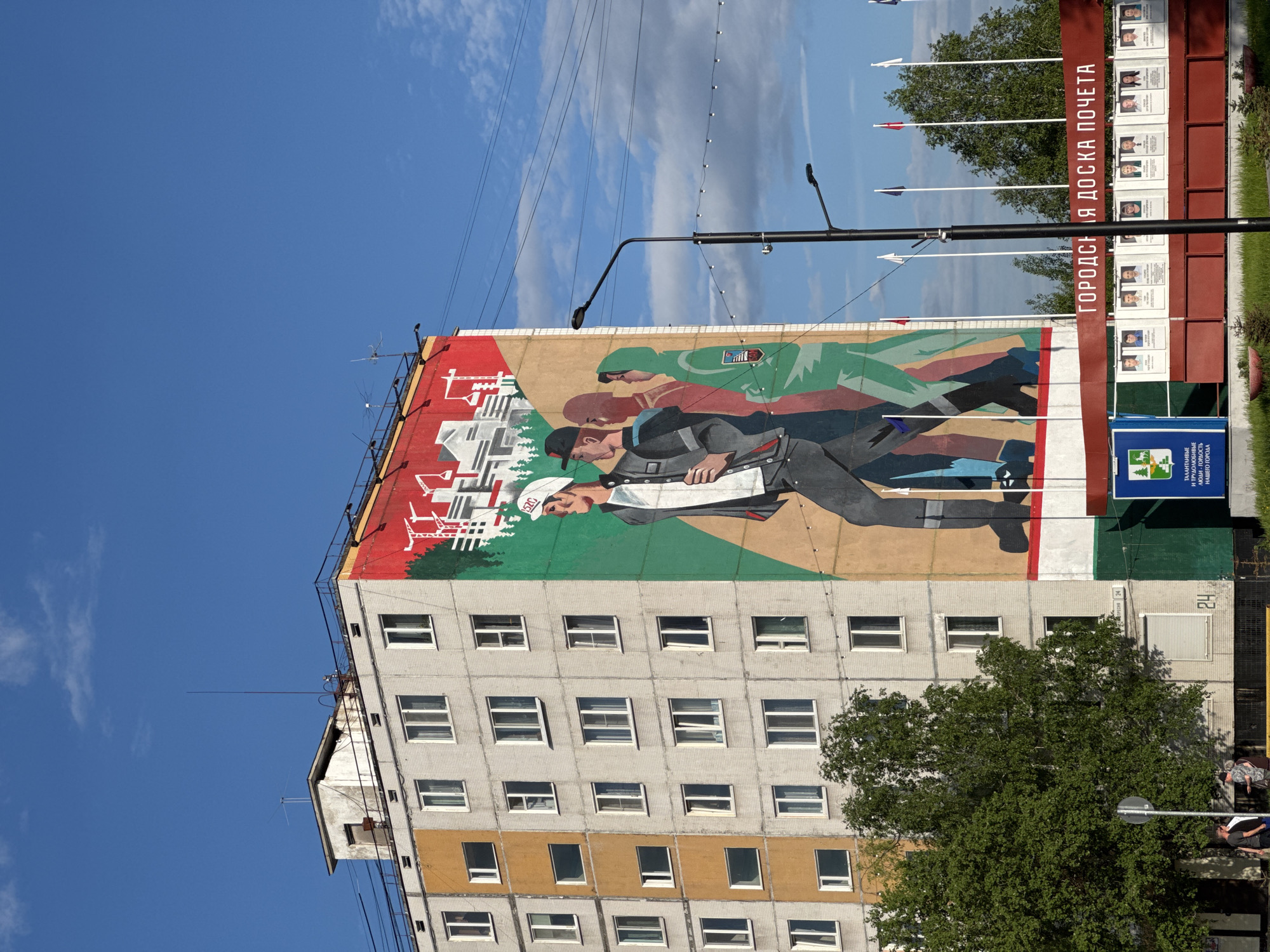 The image depicts a mural on the side of a multi-story building in Tynda, Russia. The mural features stylized figures of three people in profile, walking from right to left. The first figure, most prominent, is a male wearing a white baseball cap with a logo, a grey jacket, and dark pants. He is followed by two other figures, one in a red shirt and the other in a green hooded jacket with a crest. Behind the figures, the mural shows abstract representations of construction cranes and buildings against a green and red background.
The building itself appears to be a typical Soviet-era apartment block, constructed with grey concrete blocks and featuring numerous windows. A section of the building on the left side is painted in a yellowish-orange hue. The sky above is bright blue with scattered white clouds.
In the foreground, to the right of the mural, there is a red banner with Russian text that translates to "City Honor Board." Below this banner, a display of framed portraits of individuals is visible, suggesting it is indeed a public recognition area. There are also lampposts and a small tree. On the left side of the image, a large green tree partially obscures the building. Two people are visible standing near the base of the building on the left, and another two are further back.
The overall scene suggests an urban environment, likely a residential area in Tynda, characterized by Soviet-era architecture and public art.