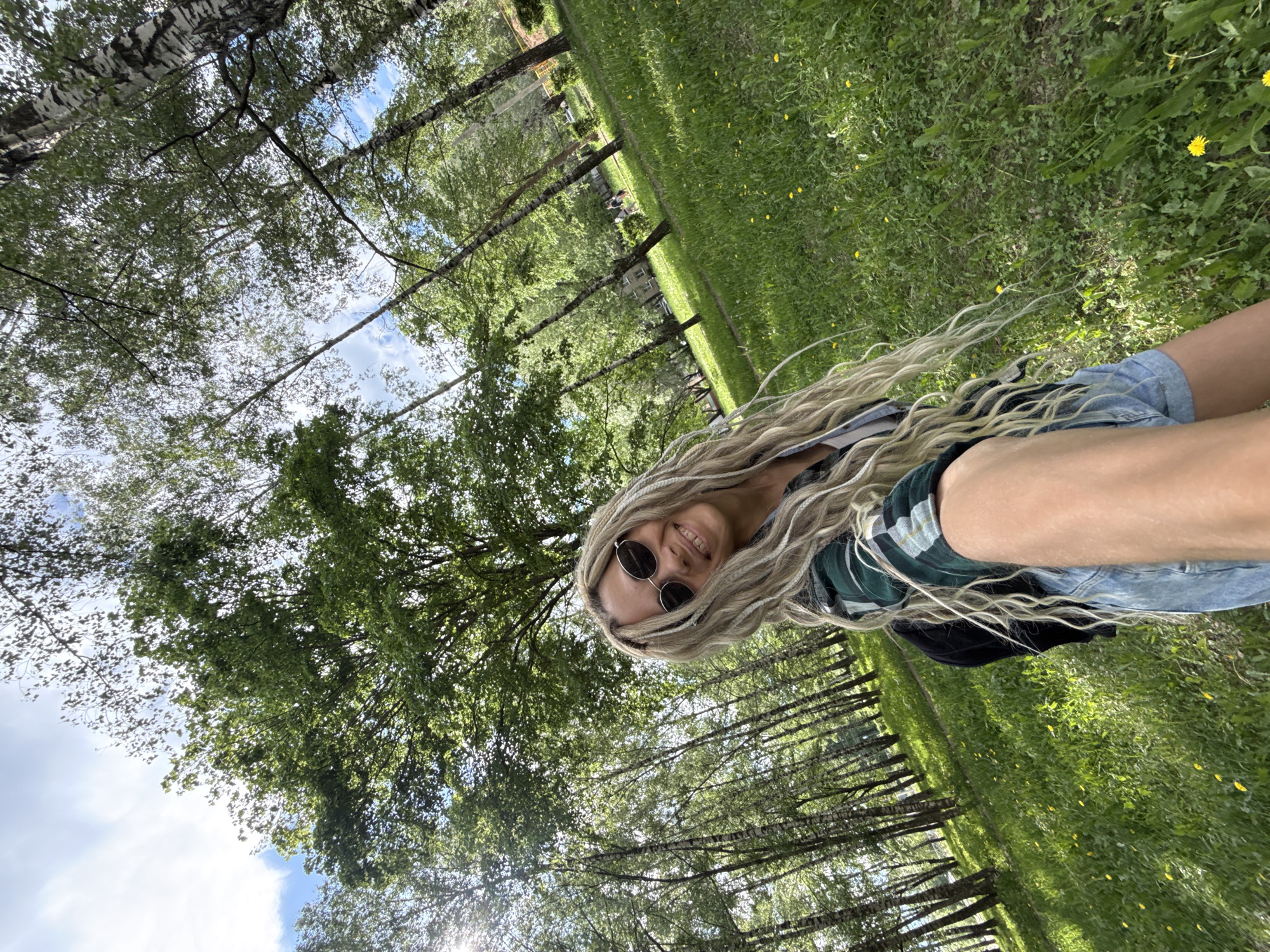 A woman with light blonde braided hair and sunglasses smiles toward the camera. She is wearing a plaid shirt and denim shorts. The photo is taken outdoors in a grassy area near birch trees. The trees form a row in the background, with more foliage visible above. Patches of sky are visible through the trees. The location appears to be a park-like setting, possibly Rusanovo, Russia, given the presence of birch trees and general landscape.