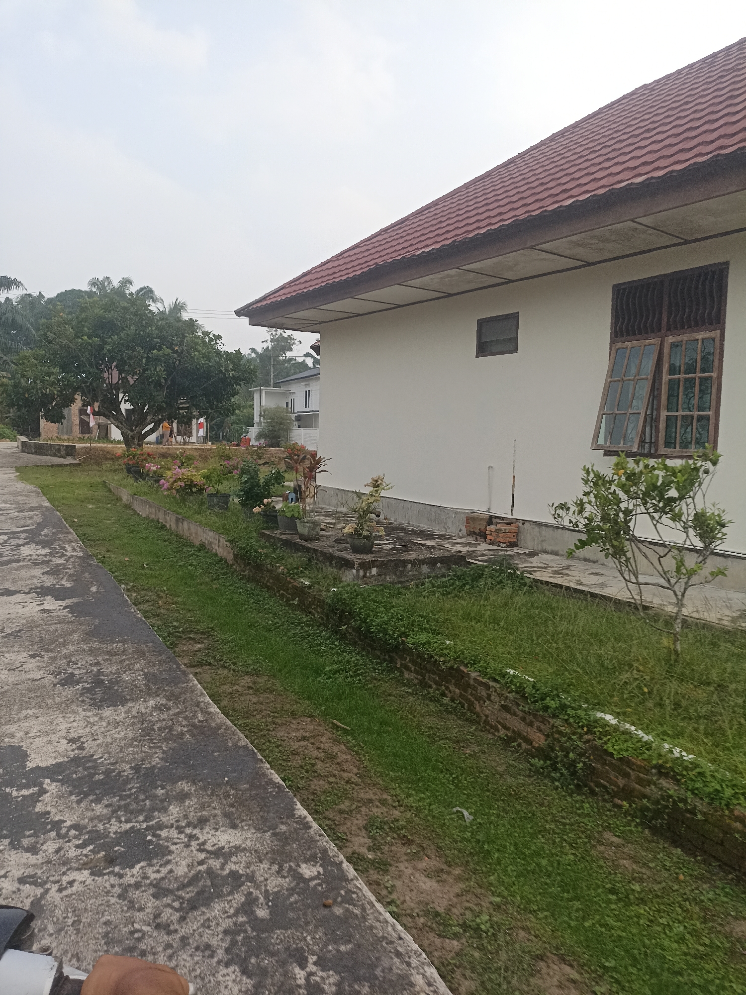 The image depicts an outdoor scene in Benteng Hulu, Indonesia, featuring a white building with a red tiled roof. The building has a large window with brown wooden shutters that are partially open, and a smaller rectangular window above it. In front of the building, a paved pathway runs alongside a strip of green grass bordered by a low brick wall. Several potted plants are placed along the pathway, with some flowering plants in the foreground. Behind the building, more trees and structures are visible in the distance. The sky is overcast, suggesting a cloudy day. The overall impression is one of a residential or institutional setting in a tropical environment.