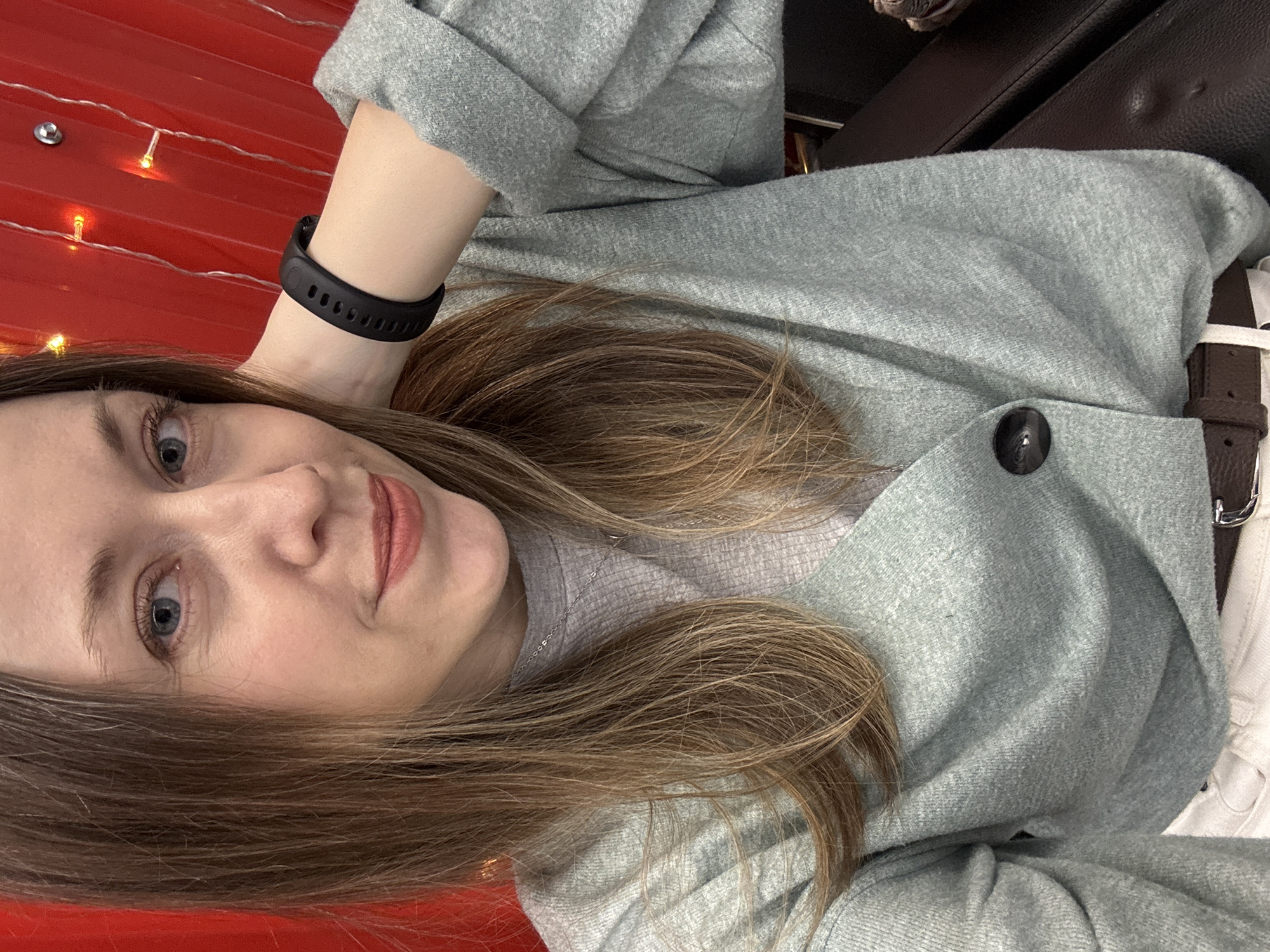 A selfie of a fair-skinned woman with light brown hair and blue eyes, likely indoors. She is wearing a gray cardigan over a gray shirt, a black fitness tracker on her wrist, and a dark brown belt with white pants. The background appears to be a red surface with string lights. The location in the city of Saint Petersburg, Russia, cannot be confirmed from the image.