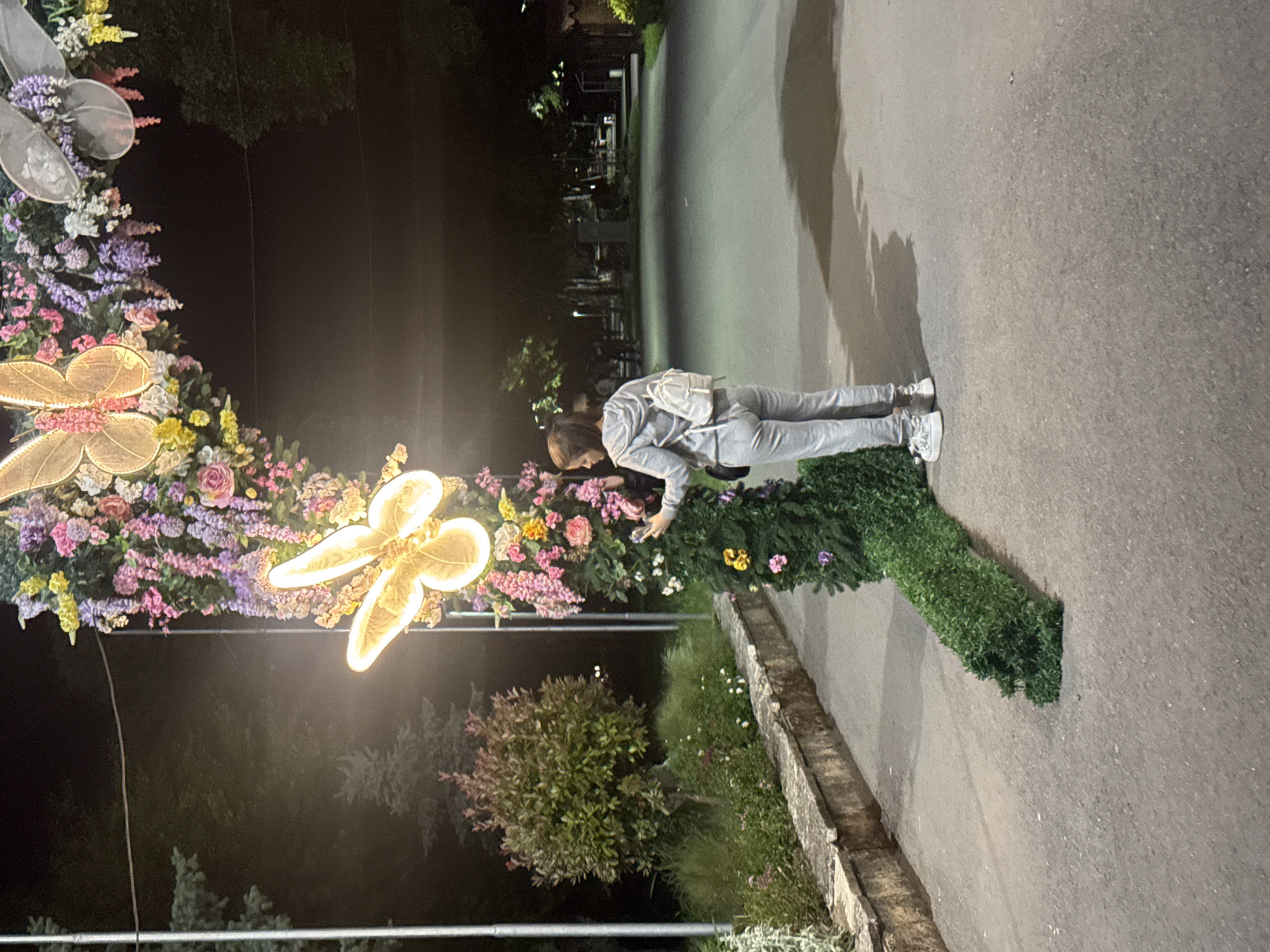 A night shot of a woman in gray sweats leaning toward a flower arrangement, in the city of Shumen, Bulgaria. The flower arrangement features a frame with a large butterfly motif and various flowers, with illuminated parts. The woman is wearing a white backpack. The background includes a paved path and some greenery, suggesting an urban park environment.