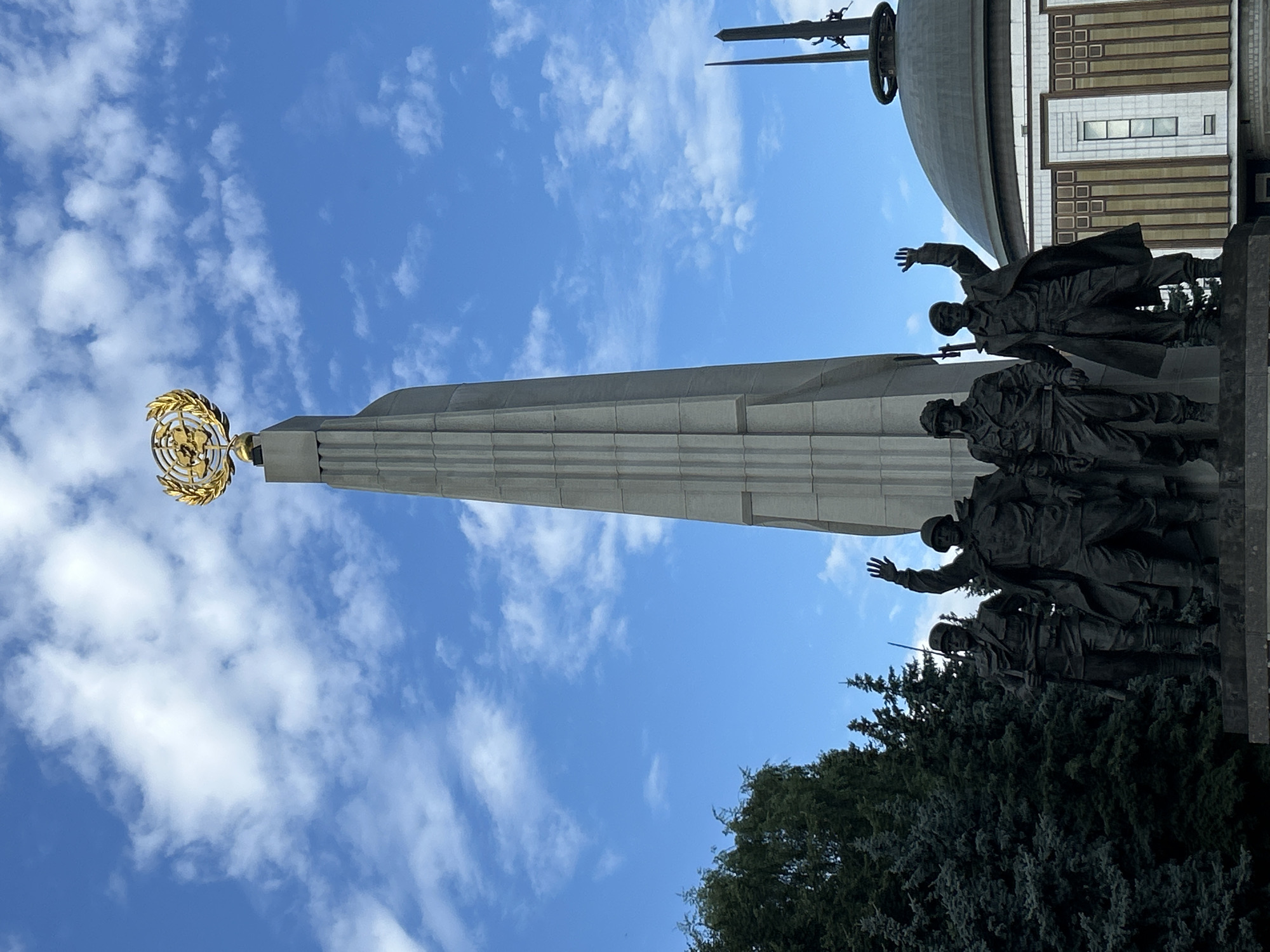 The image shows a monument with a tall obelisk topped by a golden globe surrounded by laurel leaves. At the base of the obelisk are four bronze statues of soldiers in military uniforms. To the right of the obelisk, a large domed building with a distinctive facade is visible, along with a tall, slender spire. Evergreen trees frame the left side of the monument. The scene is set against a backdrop of a bright blue sky with scattered white clouds. This monument is the Victory Monument and the domed building is the Great Patriotic War Museum, located in Moscow, Russia.