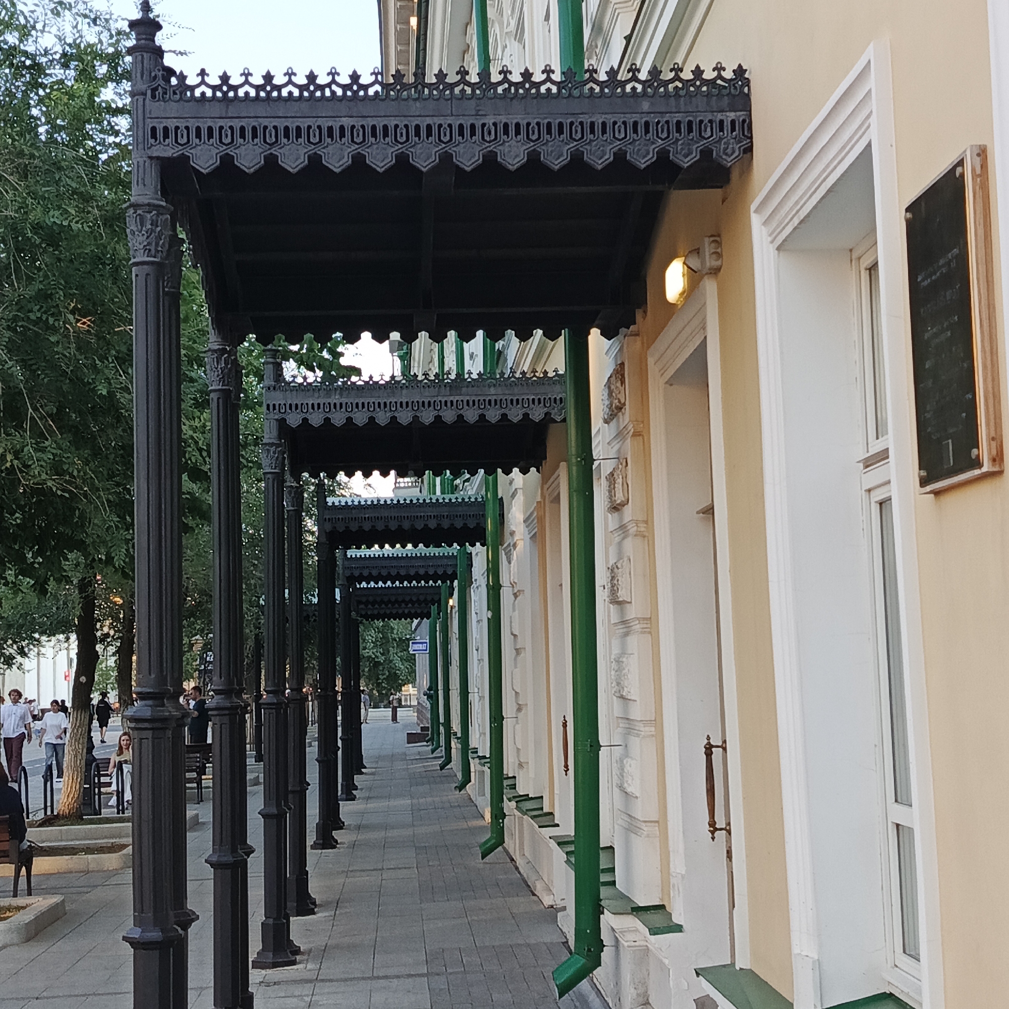 The image shows a view of a building in Orenburg, Russia, featuring a row of decorative black metal awnings supported by columns, extending along a sidewalk. The building has a light-colored facade with white-trimmed windows and green downspouts. People are visible on the sidewalk in the background, some walking and others sitting on benches. The scene appears to be in an urban setting, possibly a street or public square, with trees lining the side of the sidewalk. A plaque is affixed to the wall of the building to the right.
