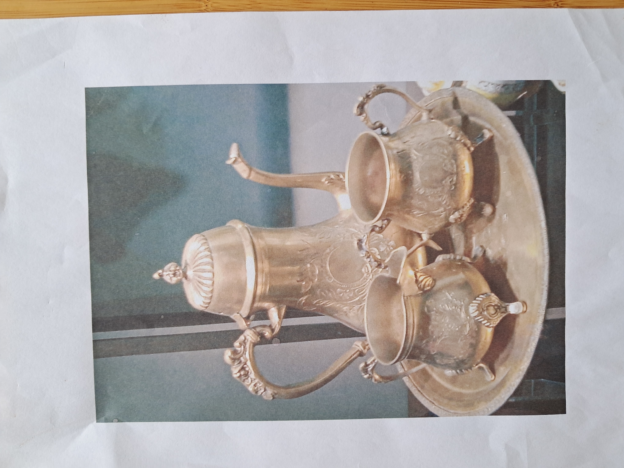 A silver tea set is displayed on a silver tray. The tea set includes a teapot with a lid, and two smaller cups or pots, one on either side of the teapot. The teapot is ornate, with decorative etching and a curved handle. The two smaller pieces also have handles and decorative etching, and each rests on three feet. The tea set is placed on a round, flat silver tray, which also appears to be etched. The background is a muted blue-grey color. The overall impression is of an antique or vintage display of silverware. The objects are arranged in a studio-like setting, with professional lighting enhancing the metallic sheen of the silver.