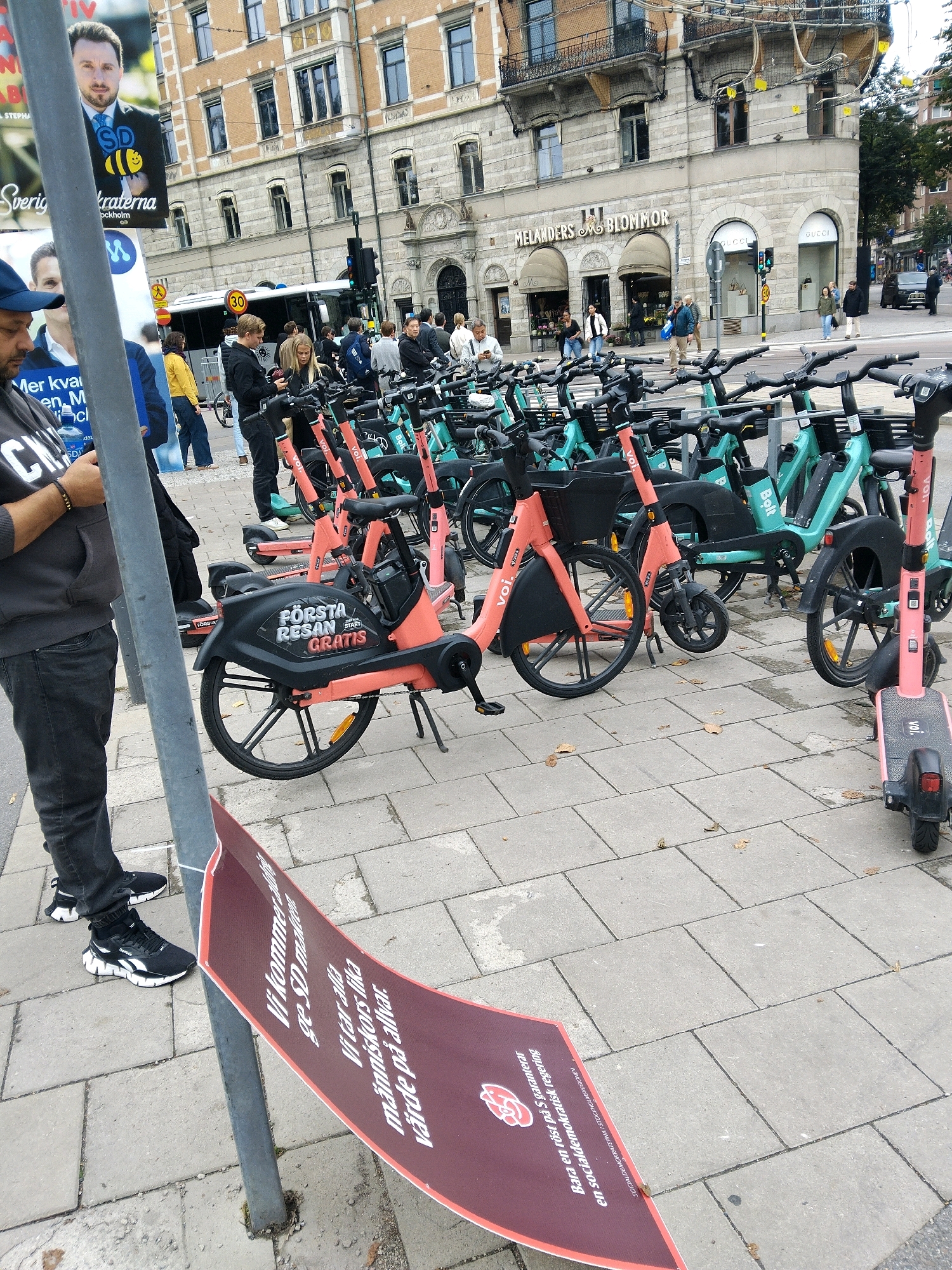 A wide-angle, eye-level, outdoor shot depicts a street scene in Stockholm, Sweden, with a prominent display of shared electric scooters and bicycles. In the foreground, a person in a dark hoodie and grey pants stands near a metal pole with a red banner. The banner has white text and a logo. Behind this, numerous shared electric scooters and bicycles are parked in rows, many of them a bright orange or teal color, with some displaying "Voi" and "Bolt" branding. The scooters and bikes are arranged on a paved sidewalk. In the background, several people are visible walking and interacting. Buildings line the street, one with signage for "MELANDERS BLOMMOR" and "GUCCI". A public bus with a "30" speed limit sign is also present. The overall environment suggests an urban commercial area.