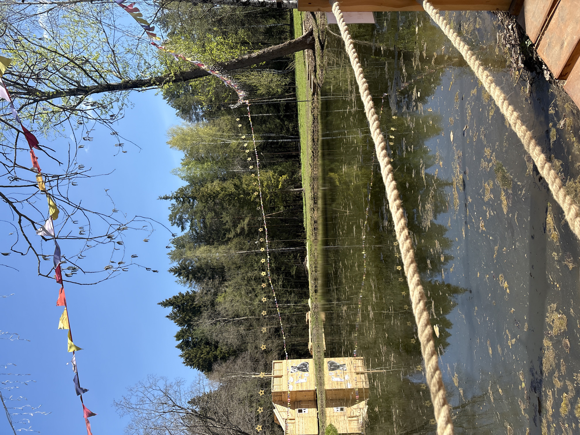 The image depicts a pond or lake in Perm, Russia, with dense trees lining the opposite shore. Strings of yellow star-shaped lights and multi-colored flags are strung above the water. A rope railing is in the foreground, extending from a wooden structure. The water's surface reflects the surrounding trees and structures, creating a mirrored effect. A small wooden building with a painted mural is partially visible on the shore.