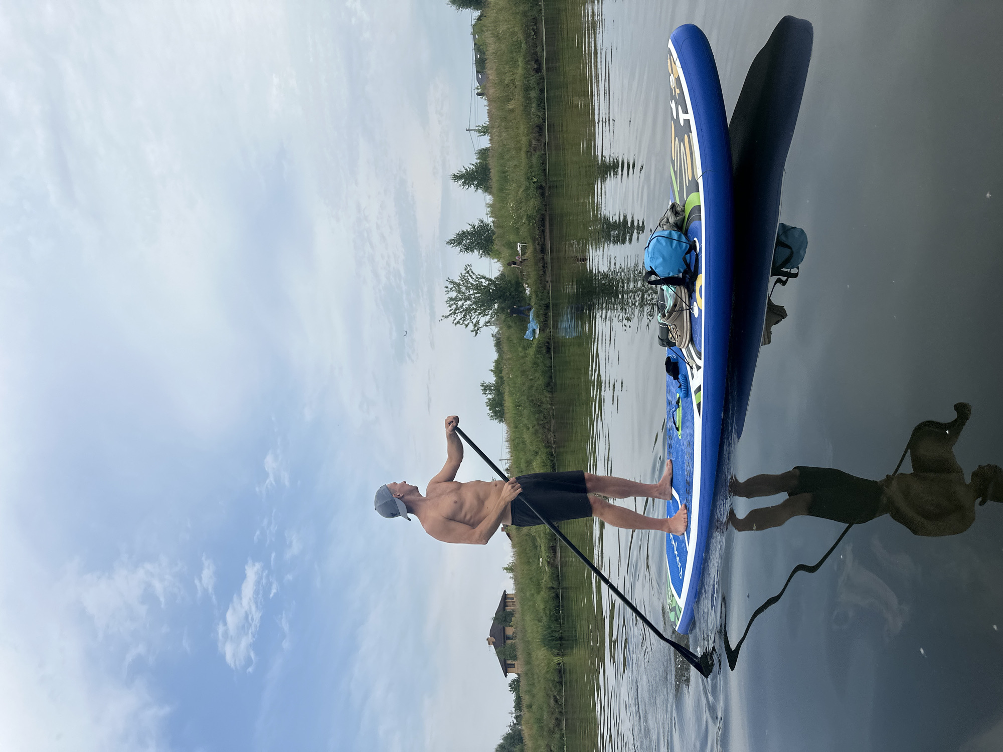 An athletic, shirtless man wearing black shorts and a grey baseball cap is stand-up paddleboarding on a calm body of water, likely a river or lake, in the city of Gorki Leninskiye, Russia. He is holding a paddle, with the blade submerged in the water, and looking to his right. His reflection is visible in the still water. The paddleboard is blue with colorful graphics. On the front of the paddleboard, there are two bags and a pair of shoes. In the background, a grassy embankment lines the water, with trees and a building visible. The sky is cloudy with patches of blue.