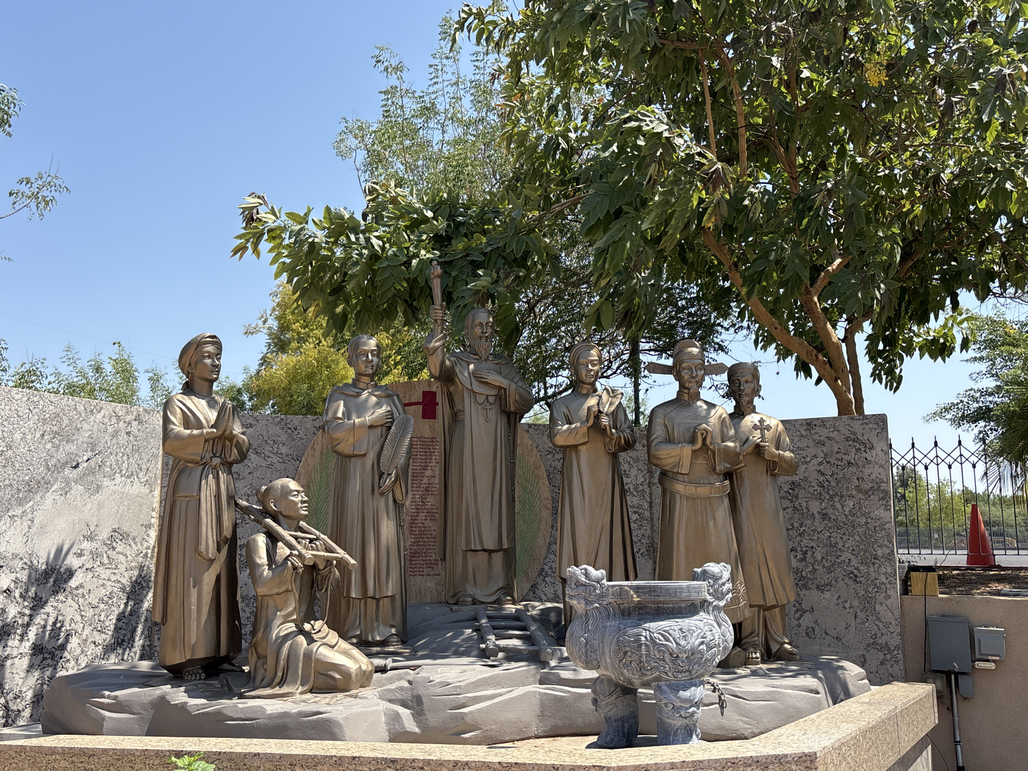 The image depicts a bronze statue of several individuals, likely representing religious figures, in an outdoor setting. The statues are arranged in a group, with some figures standing and one kneeling. One prominent figure in the center holds a torch. Behind the statues is a stone wall with what appears to be an inscription. In the foreground, there is a large, ornate stone urn. The scene is set against a backdrop of trees and a clear blue sky, suggesting a sunny day. The statues appear to be part of a monument or memorial. The question indicates this is in Phoenix, Arizona, United States.