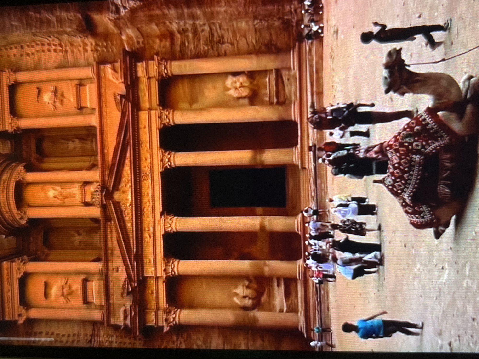 The image shows Al-Khazneh ("The Treasury") in Petra, Jordan. The ornate facade of the rock-cut building is visible, carved into the sandstone cliffs. In the foreground, there is a crowd of tourists and a camel with a decorated saddle blanket. People are walking around the site, exploring the historical landmark. The ground appears sandy and uneven. The image is rotated 90 degrees counter-clockwise. Tinaquillo, Venezuela is not depicted in the image.