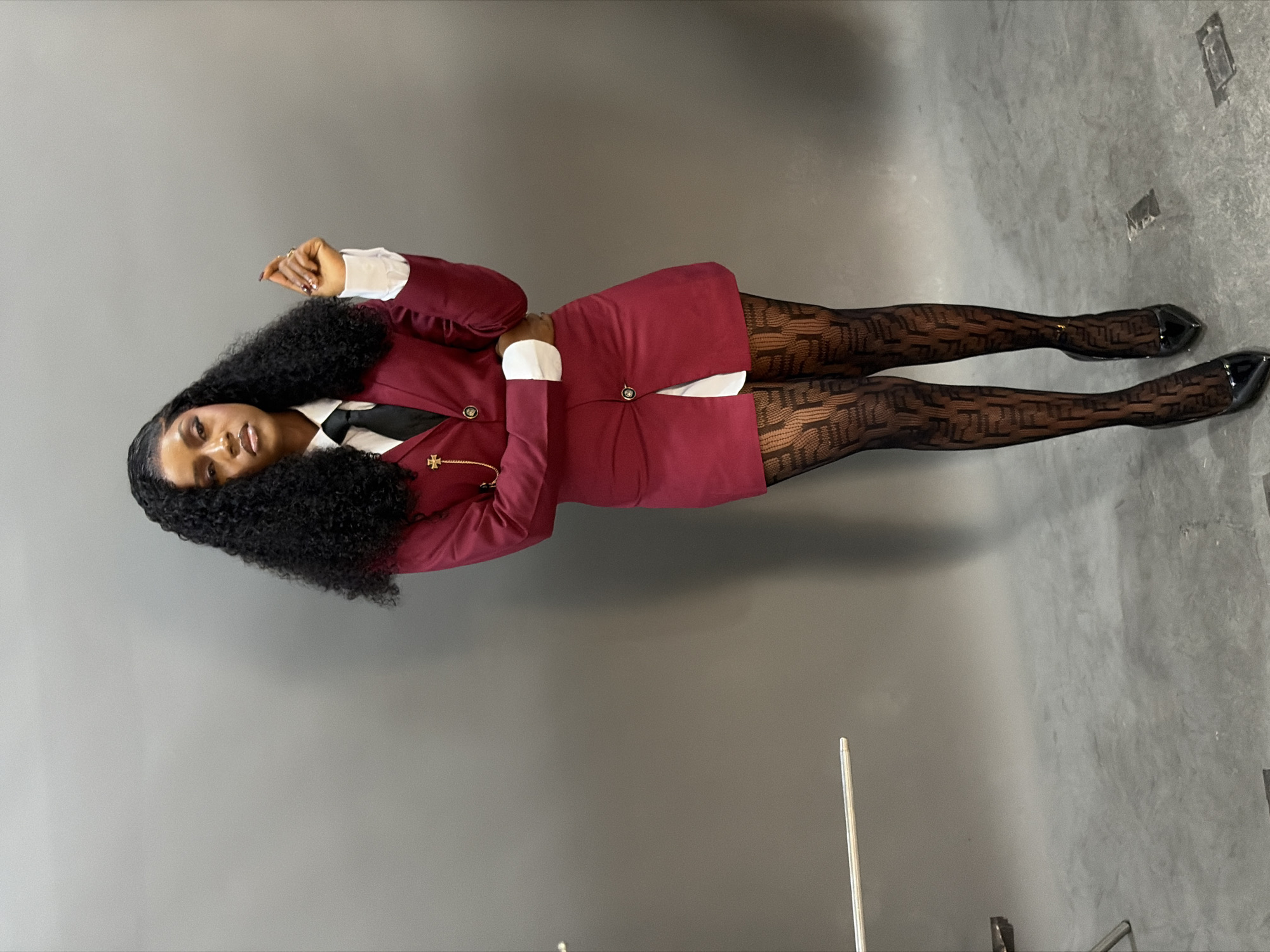 A female subject is captured in a full-body studio portrait against a plain grey backdrop. She stands with her left hand raised and fingers slightly curled, while her right arm is bent across her midsection. Her dark, voluminous curly hair falls past her shoulders. She wears a white collared shirt, a black tie, and a fitted burgundy blazer. The blazer features a single gold-colored button closure, white cuffs, and a decorative gold-colored chain with a cross-like emblem pinned to the left lapel. She also wears a matching burgundy mini-skirt with a gold-colored button detail. Her legs are clad in sheer black stockings patterned with repeating 'F' letter designs. Black patent leather pointed-toe high heels complete her attire. The studio floor is a darker shade of grey, showing some textured wear and faint rectangular markings. Partially visible on the left side of the frame are a silver metal pole and the base of a stand, indicative of studio equipment.