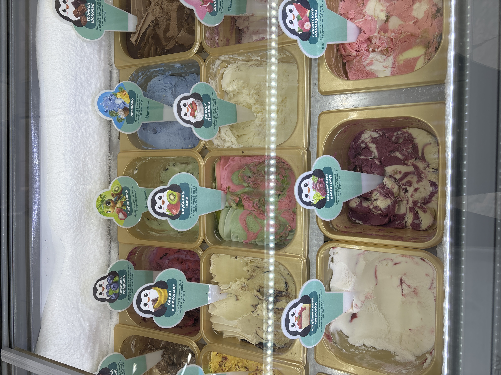 The image captures a vibrant display of various ice cream and sorbet flavors within a commercial freezer, likely in a shop or cafe in Chaikovsky, Russia. The freezer is illuminated by a vertical strip of LED lights on the right side, casting a bright glow over the tubs of frozen treats. A thick layer of white frost or ice is visible along the left interior wall of the freezer.
The ice cream and sorbet are neatly arranged in multiple rectangular containers. Each container features a descriptive label shaped like a spoon or shovel, denoting the flavor. Most labels feature a cartoon penguin mascot, while others include different cartoon characters like a hippo for "Лимпопо" (Limpompo) and a turtle for "черепашка" (turtle), along with the brand name "33 пингвина" (33 penguins).
A variety of flavors and colors are visible, including:
- "тройной шоколад" (triple chocolate): A rich brown ice cream.
- "Десерт Павлова" (Pavlova dessert): A creamy, pale yellow ice cream.
- "гавайские каникулы" (Hawaiian holidays): Pink and white swirled ice cream.
- "испанский виноград" (Spanish grape): A distinctive purple and white swirled ice cream.
- "клубника-киви" (strawberry-kiwi): Features pink and green swirls.
- "клубничный чизкейк" (strawberry cheesecake): White ice cream with red swirls.
- "банан в шоколаде" (banana in chocolate): A creamy, pale yellow ice cream.
- "черника-голубика" (blueberry-lingonberry): A dark reddish-purple ice cream.
- "Лимпопо" (Limpompo): A light blue ice cream.
- "черепашка" (turtle): A bright green and yellow sorbet.
The labels provide specific descriptions, such as "Пломбир с молочным и темным шоколадом и шоколадным кремом" (Plombir with milk and dark chocolate and chocolate cream) for triple chocolate, or "Пломбир со вкусом нежного безе и клубничным декором" (Plombir with tender meringue flavor and strawberry decor) for Pavlova dessert. The scene highlights a wide selection of visually appealing frozen desserts.