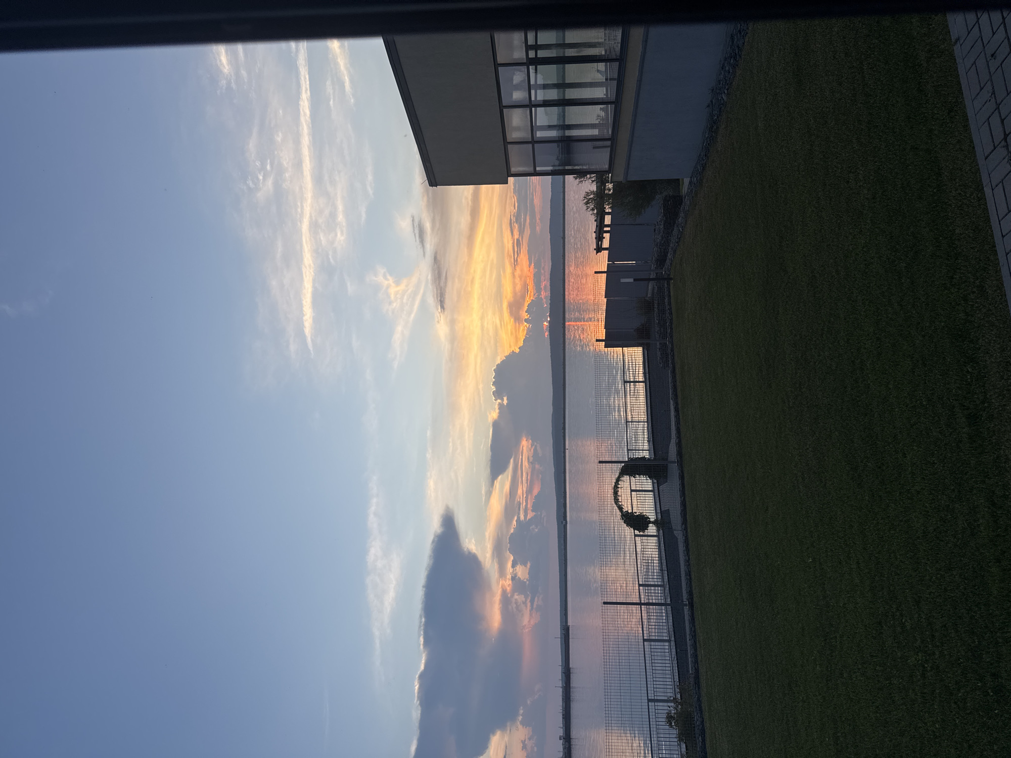 This image shows a sunset over the Kama River in Chaikovsky, Russia. The sky is filled with soft hues of orange, pink, and blue, with wispy clouds catching the last rays of the sun. In the foreground, a well-maintained lawn leads to a fence and a walkway. A modern building with large windows is visible on the right side of the frame. The river reflects the colors of the sky, creating a serene and picturesque scene. There are no people visible in the image.