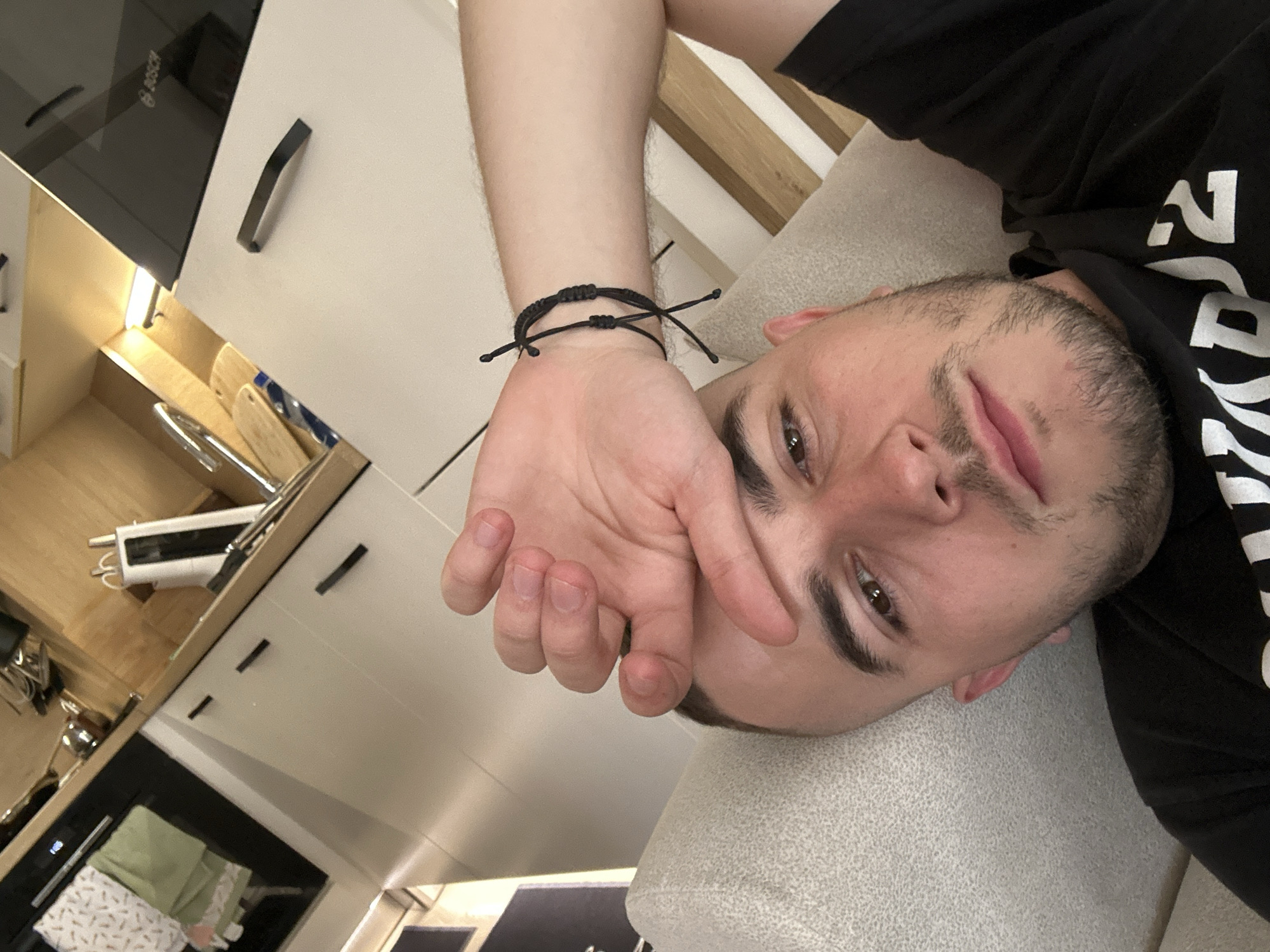 A man lies on a light gray textile surface, likely a sofa or bed. He wears a black t-shirt with white lettering and a black bracelet on his left wrist. His left hand is raised toward his face, near his forehead. He has short brown hair and a beard. The background includes white cabinets and drawers with black handles, and a black Bosch oven or stovetop. Based on the presence of household appliances and furniture, the location is most likely inside an apartment or home, possibly in Plovdiv, Bulgaria. No specific landmark or identifiable feature indicates a precise location within the city.