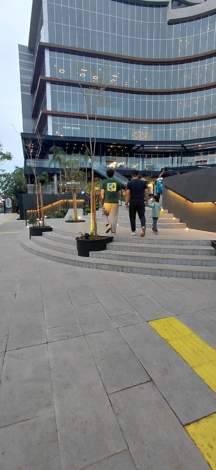 The image depicts a modern architectural building with a curved facade, featuring large windows with dark frames. In the foreground, a set of wide, curved concrete stairs ascends towards the building. The stairs are illuminated by subtle uplighting along the edge of the landings. A paved walkway with large, grey rectangular tiles leads to the stairs, with a distinct yellow tactile paving strip on the left side.
Three individuals are walking up the stairs away from the camera. The person in the middle wears a green t-shirt with a graphic and light-colored pants. To their right, a person in a dark t-shirt and dark pants is walking, holding the hand of a child wearing a light blue jacket and grey pants.
Several small trees in dark circular planters are strategically placed along the stairs and walkways, adding greenery to the urban environment. The building's lower levels appear to house commercial spaces, with signage visible, including "SPORT HOUSE" and "MARTINEZ". The overall scene suggests an outdoor public space in a city, likely a commercial or entertainment complex.
This scene is located at the Plaza de la Costanera, a prominent public space in Encarnación, Paraguay, known for its modern design and waterfront location.