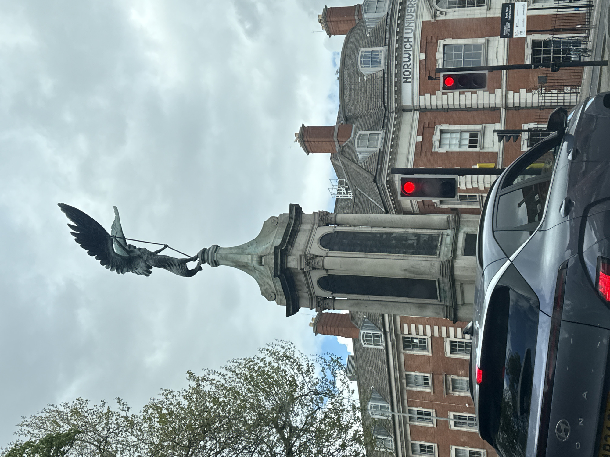 The image captures a street scene in Norwich, United Kingdom, featuring the Norwich War Memorial. The monument, topped by a bronze statue of a winged angel holding an olive branch, stands tall beside the road. The background includes a portion of the Norwich University of the Arts building, characterized by its red brick facade and gray roof. Traffic lights with illuminated red signals are visible. To the right, a dark-colored Hyundai Kona car occupies a significant portion of the frame. The sky is overcast with gray clouds, and bare branches of a tree are visible at the bottom left corner.