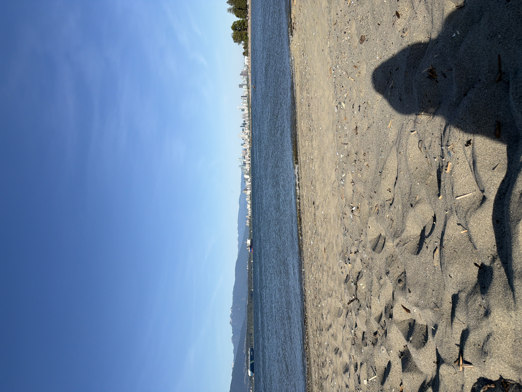 A long horizontal shot of a beach with a shadow on the right side and the ocean on the left. The sky is blue and clear. In the distance on the other side of the water, there is a mountain range, and a city skyline is barely visible. A forensic analysis would indicate that the location is likely a beach in Vancouver, Canada, given the topography and cityscape.