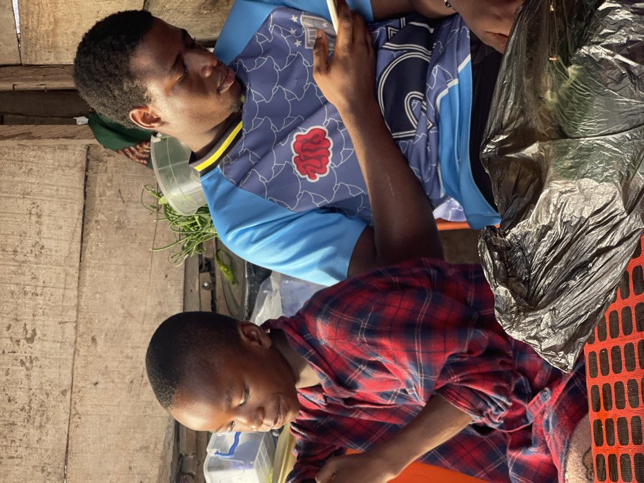Two young men are visible. One wears a blue soccer jersey with a red design and yellow collar, looking at his phone. The other is wearing a red and blue flannel shirt and is looking down at a container. A black plastic bag sits on the right side. The scene is outdoors with wooden walls in the background and various containers and plants. The image is tilted to the side.