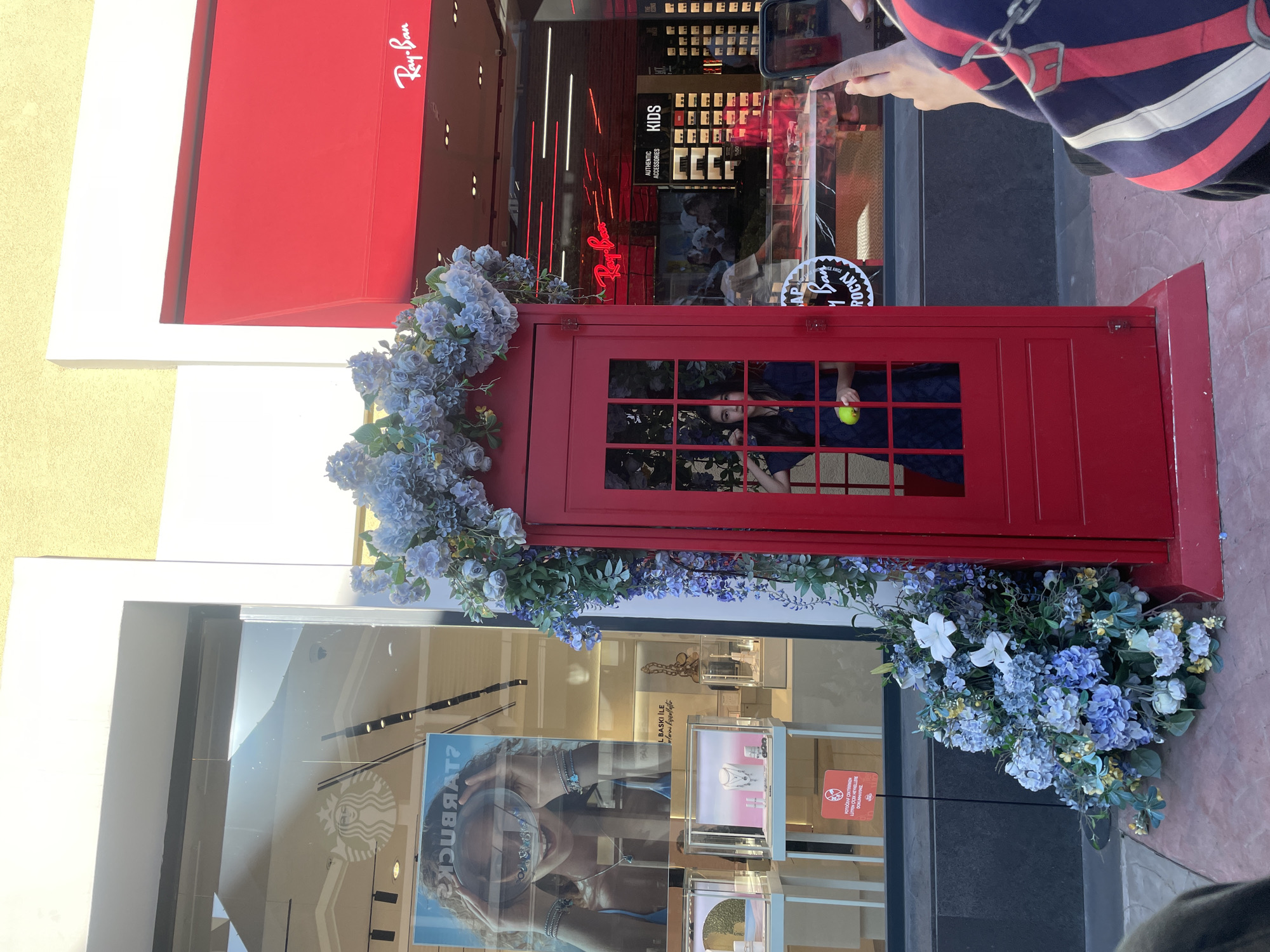 A person is taking a photo of a red phone booth decorated with blue flowers. The phone booth is in front of a store with a red awning and a Ray-Ban logo. Behind the phone booth, a young girl is visible inside, holding a yellow object. The scene appears to be an outdoor shopping area.