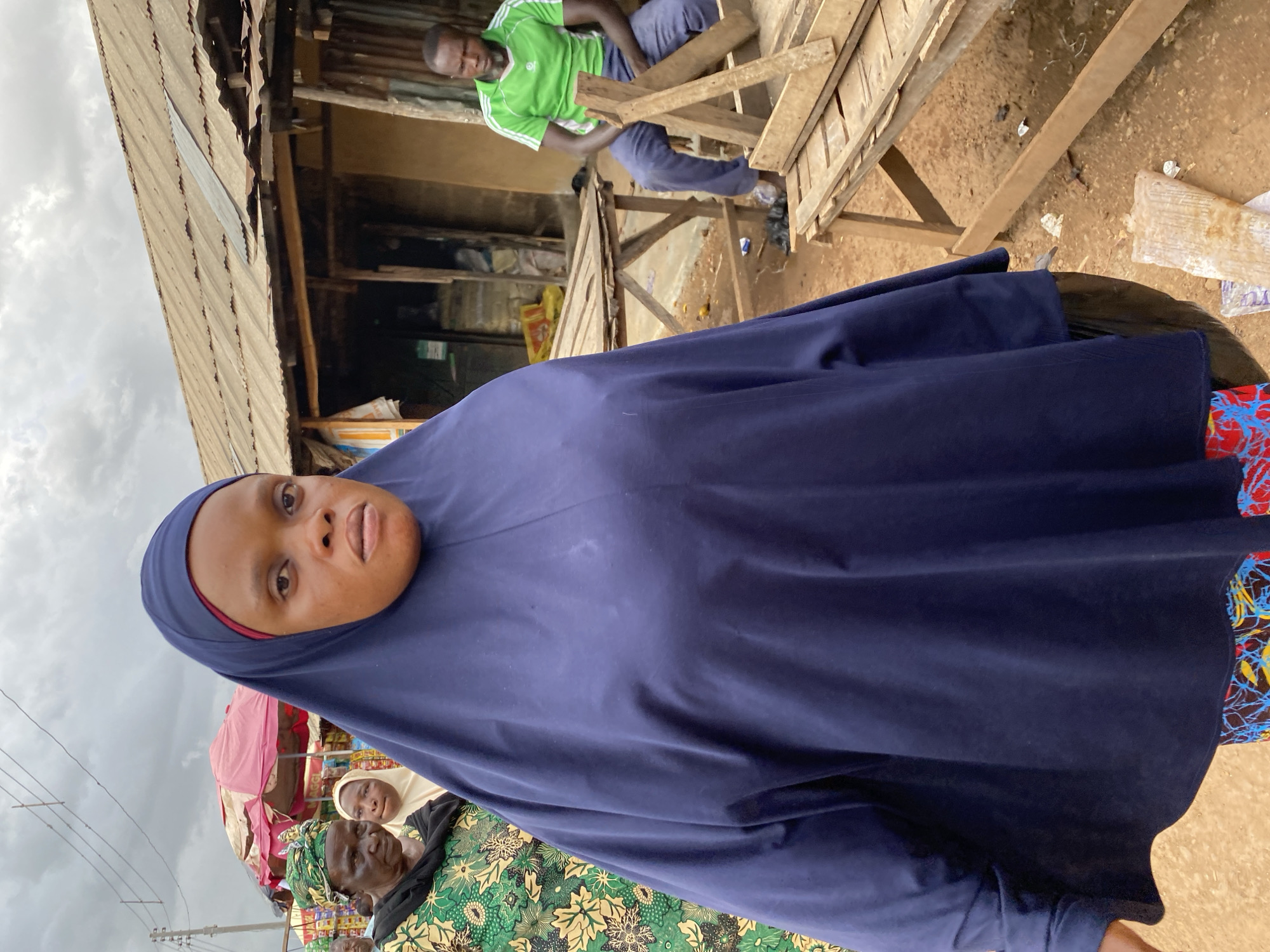 A medium shot captures a woman wearing a dark blue hijab and a patterned skirt, standing in what appears to be an outdoor market in Bukuru, Nigeria. She is positioned in the foreground, with her face turned slightly towards the camera. To her left, two older women are visible, one wearing a green and yellow patterned outfit and a headscarf, and another partially obscured behind her. In the background, a man in a green t-shirt and blue pants sits on wooden structures, possibly a market stall. Several wooden tables and benches are scattered throughout the scene, and a corrugated metal roof structure is visible behind the man. The ground is dirt, with some debris present. The sky is overcast.