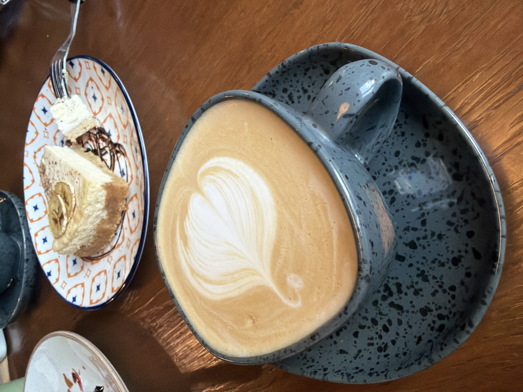 A close-up, high-angle shot captures a grey speckled coffee cup filled with latte art, placed on a matching saucer, set on a wooden table. The latte art features a white heart shape created with milk foam. To the upper right, a slice of cheesecake with caramelized banana slices and chocolate drizzle sits on a plate with an orange and blue geometric pattern. A fork with a small piece of cheesecake on it is poised above the slice. The background is blurred, suggesting an indoor setting, likely a cafe or restaurant. The image does not contain information to identify its location in Buzuluk, Russia.
