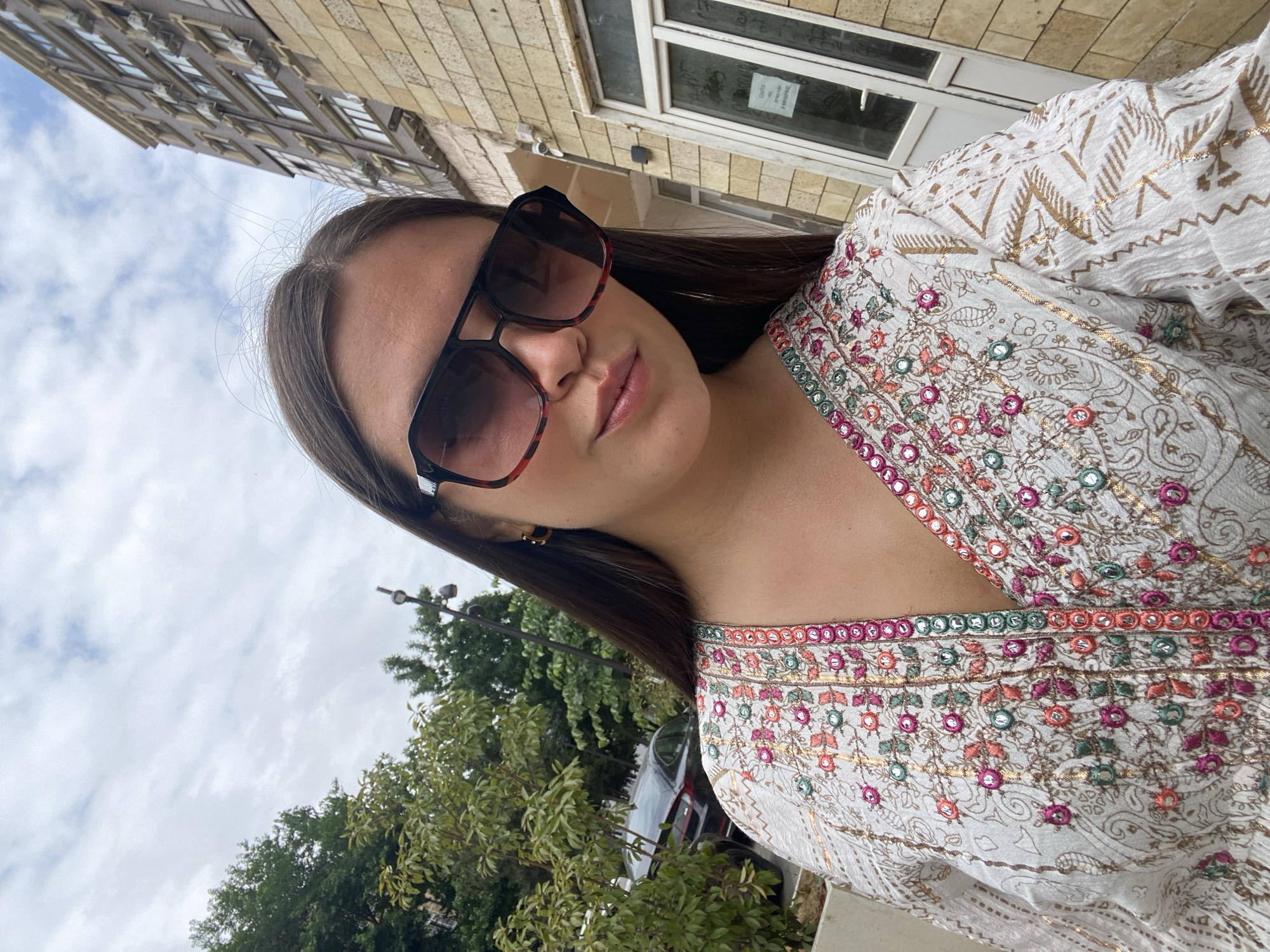 A young woman is taking a selfie outdoors in front of a building. She has long, dark hair and is wearing large, dark sunglasses with red accents on the frames and a white, embroidered top. The background shows a multi-story building with many windows and air conditioning units, as well as some trees and a parked car. The sky is overcast. The woman's attire and the architecture suggest the location is likely in Derbent, Russia, a city known for its historical significance and proximity to the Caspian Sea.