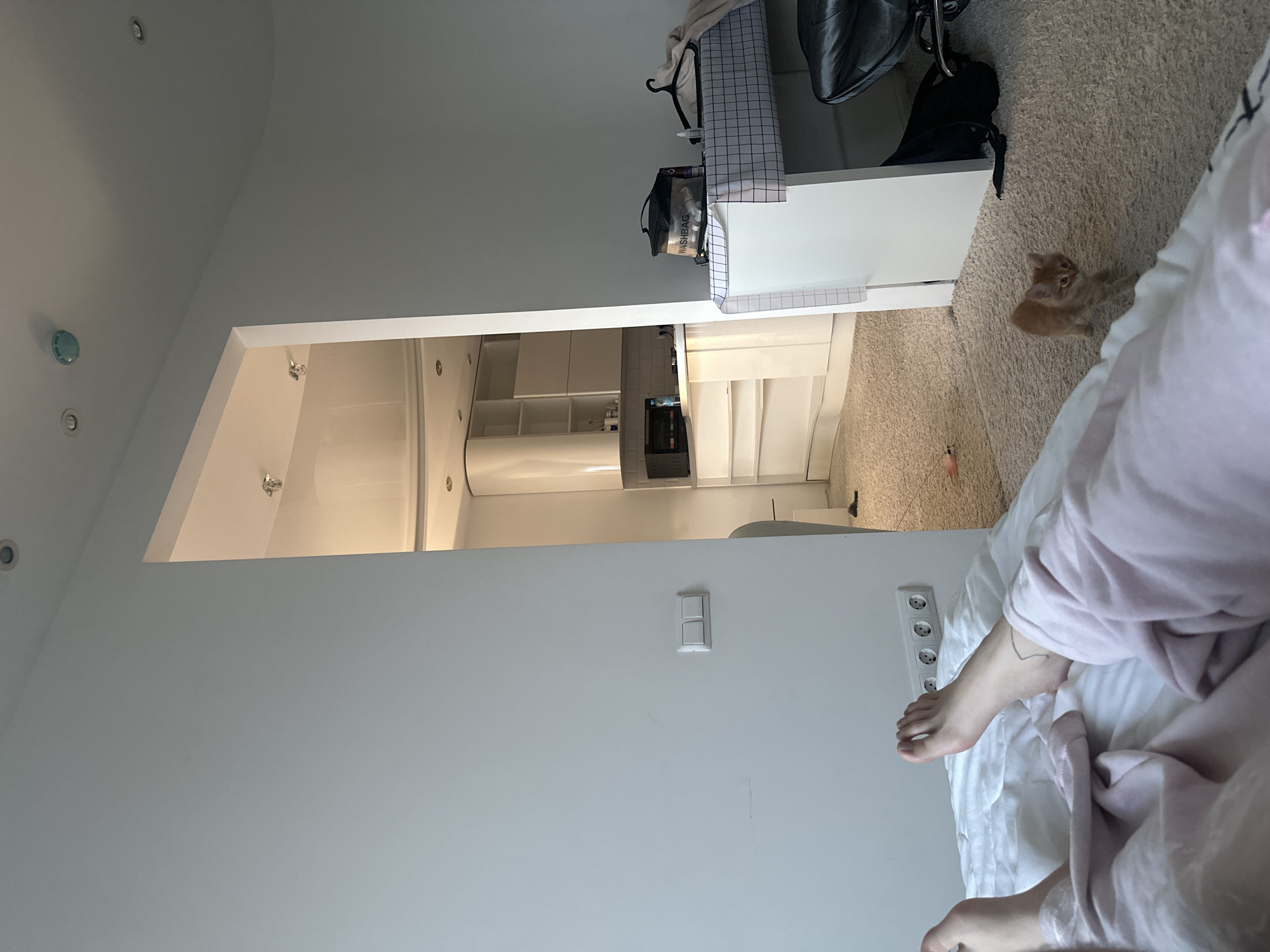 The image presents an indoor scene captured from a low angle, suggesting a perspective from someone lying in bed. In the immediate foreground, a person's bare feet are visible, resting on white bedding and partially covered by light pink fabric. A light gray wall runs along this side of the frame, featuring a white triple power socket and, further along, a white double light switch.
The view opens into an adjacent room through a wide, white archway. This background area appears to be a modern kitchen or kitchenette, furnished with sleek, white cabinetry, some of which are open, revealing storage. An integrated microwave or oven is visible, and under-cabinet lighting illuminates the space. Recessed spotlights are embedded in the ceilings of both rooms, and a light blue-green smoke detector or sensor is mounted on the ceiling of the foreground room.
To the right of the archway, within the foreground room, a white desk or cabinet is positioned. On its surface, a black bag with the text "WASHBAG" is visible, alongside other items. A black bag and a black office chair are situated near this furniture piece. The desk surface is covered with a white and gray checkered pattern.
The floor throughout the visible area is covered by a light beige, textured carpet. A small, light orange kitten is seated on this carpet near the desk, looking towards the camera. A small toy with red or pink feathers lies on the carpet close to the kitten. The overall impression is one of a calm, domestic interior.