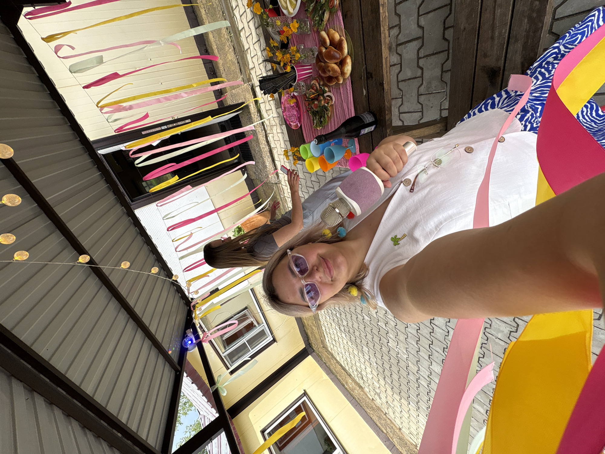 The image captures an outdoor gathering under a covered patio, likely in Birobidzhan, Russia, given the context. A woman in the foreground, wearing sunglasses and a white top, is holding a pink microphone and taking a selfie. She has blonde hair and is adorned with colorful earrings. To her left, a table is laden with party food, including pastries, salads, and yellow flowers in vases. Several colorful plastic cups and a bottle of wine are also present on the table. In the background, another woman with long brown hair stands near a doorway, and a yellow building with windows is visible. The patio is decorated with strings of lights and colorful ribbons that hang down. The ground is paved with interlocking concrete stones.