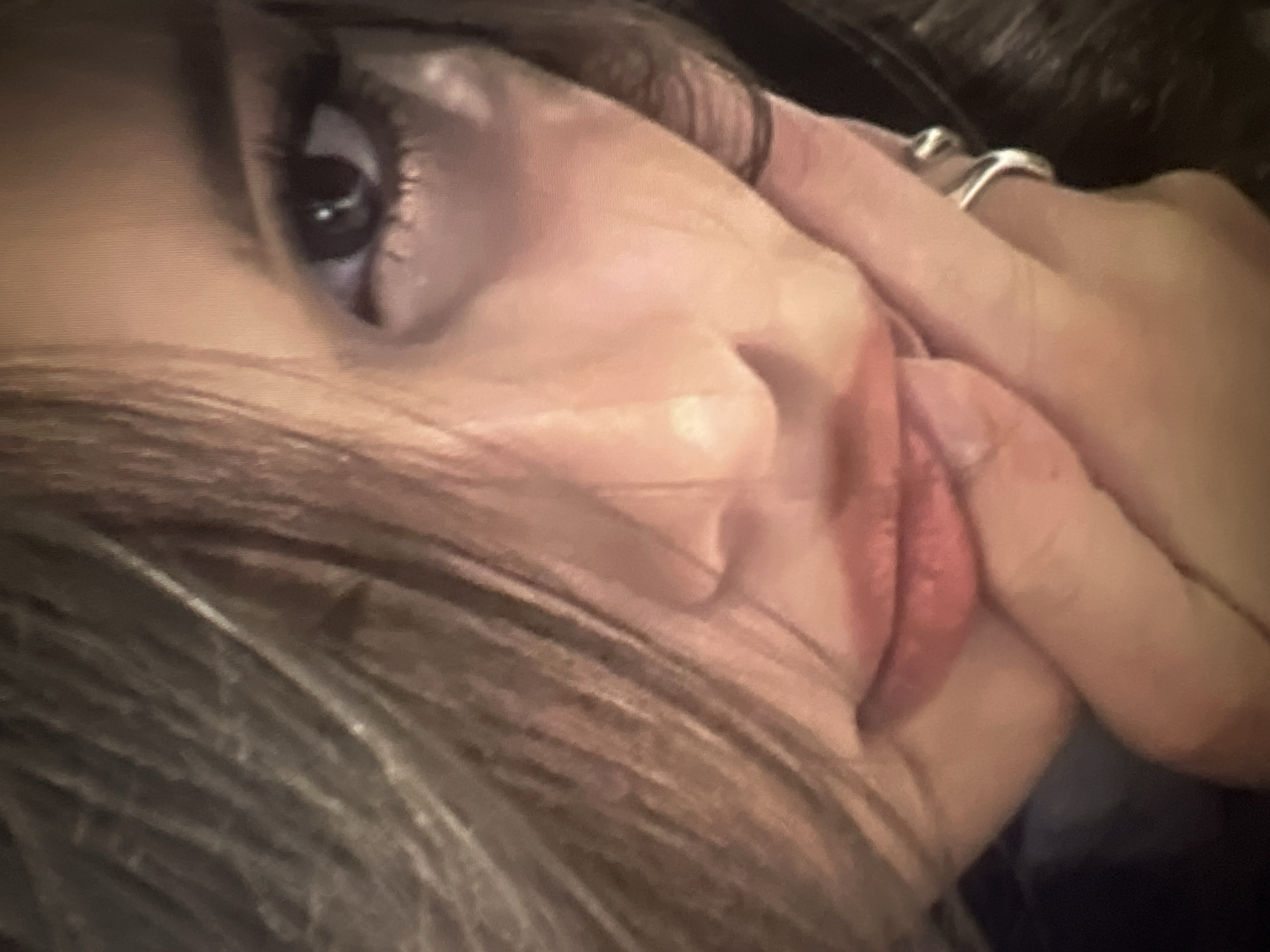 A close-up shot focuses on the right side of a woman's face. She has brown hair that partially obscures her features, particularly her left eye. Her right eye is open and appears to be looking towards the camera, with dark eyeliner and mascara visible. Her lips are slightly parted, revealing a hint of her teeth. Her right hand is resting on her chin, with a silver ring on her index finger. The background is dark and out of focus, making it difficult to discern any environmental details. There is no information in the image to determine the location of the city administration of Qarağandı, Kazakhstan.
