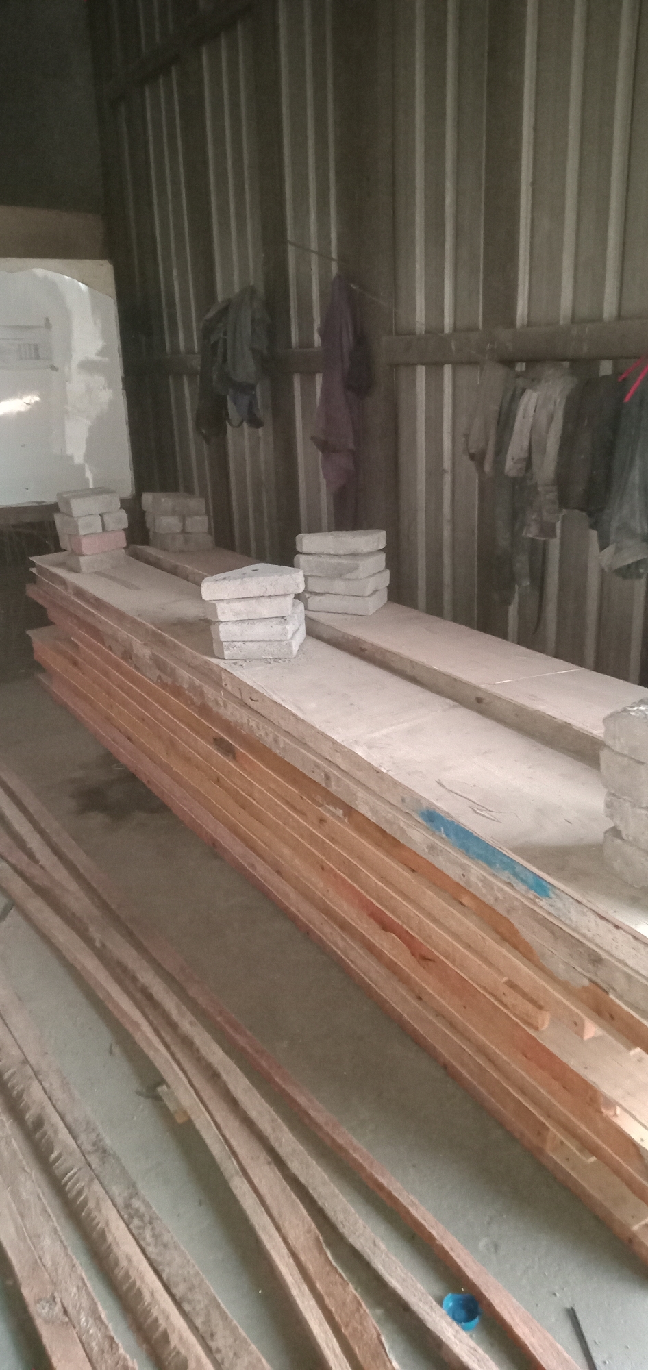 The image depicts an interior space in Sumput, Indonesia. The primary features are stacks of lumber and concrete blocks. Some of the lumber is stacked on top of other wider pieces. Several stacks of concrete blocks are placed atop these wider pieces, seemingly for support or storage. In the background, a corrugated metal wall is visible, with items such as clothing hanging from it. A white board can also be seen in the back corner. The floor is concrete. There are no people visible in the image.
